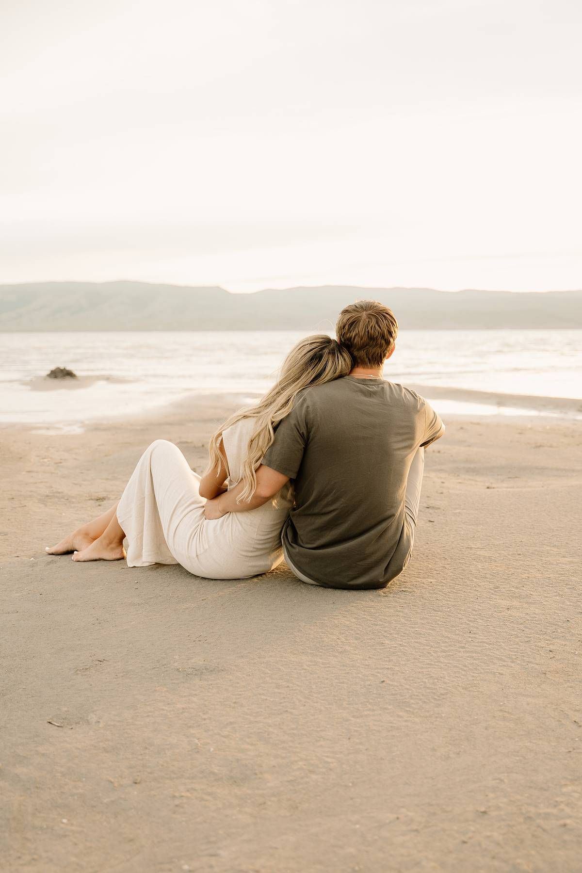 A couple sits close together on a sandy beach at sunset overlooking calm waters and distant hills.