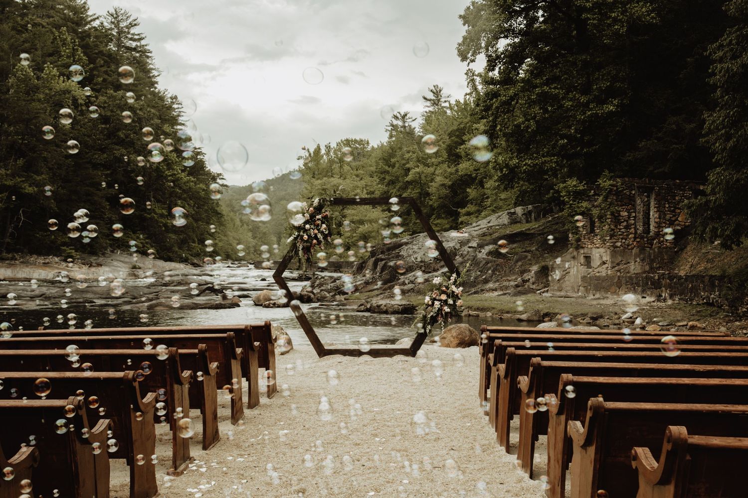 Wedding ceremony setup with wooden pews under trees and floating bubbles in the air.