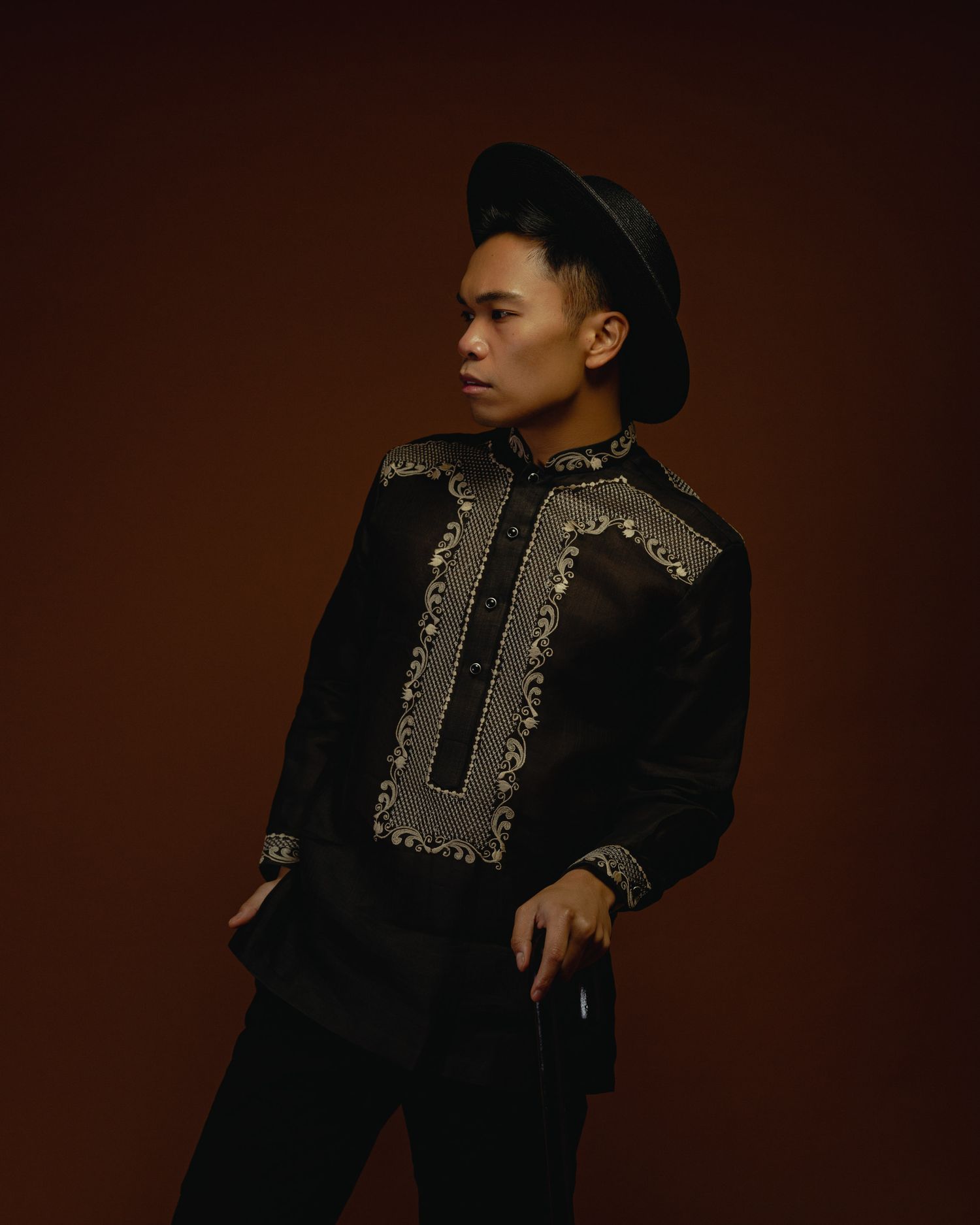 A person wearing a black embroidered jacket and cap poses in a dramatic portrait against a dark brown backdrop.