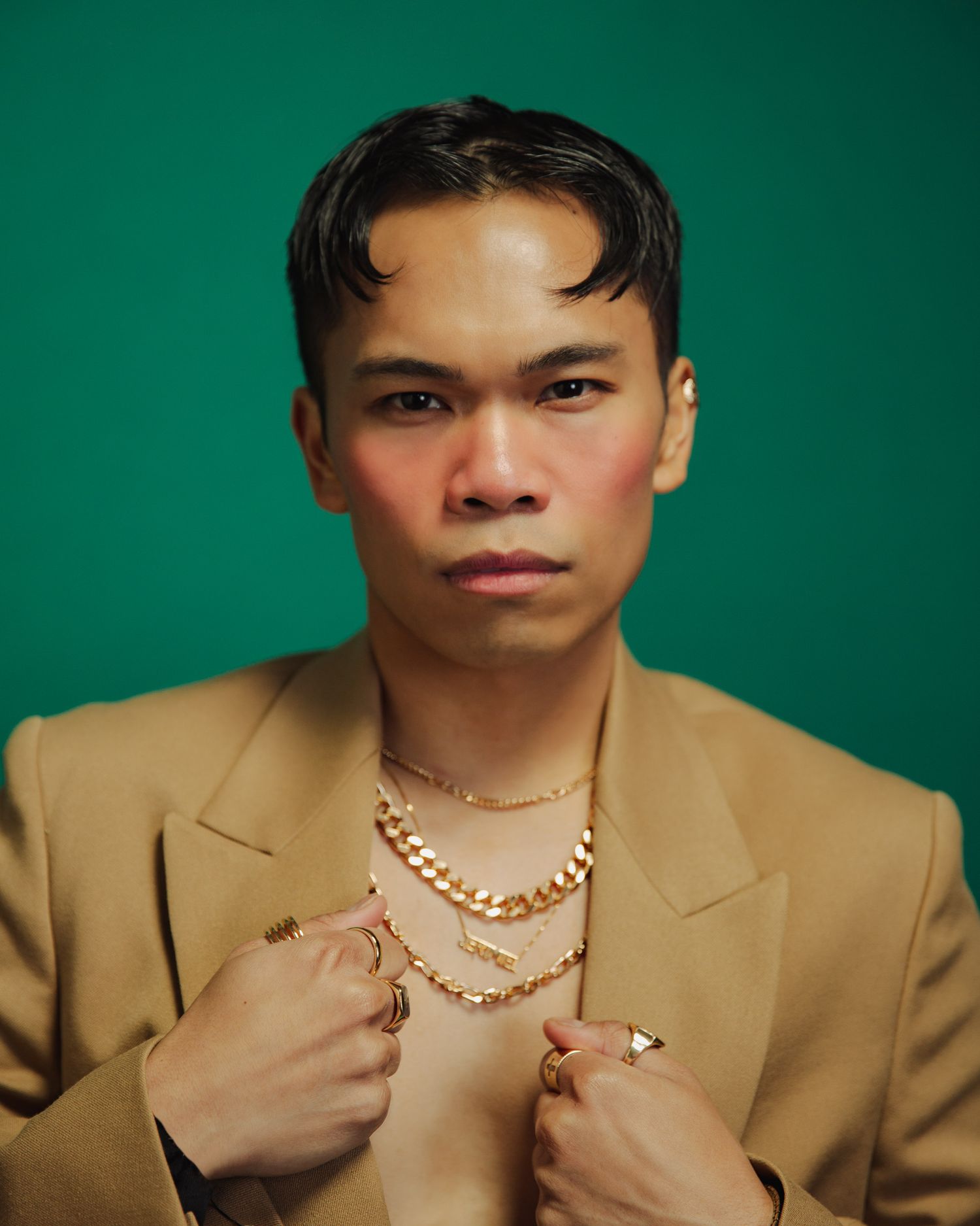 Portrait against green background shows person in tan blazer with layered gold necklaces and bare chest beneath.