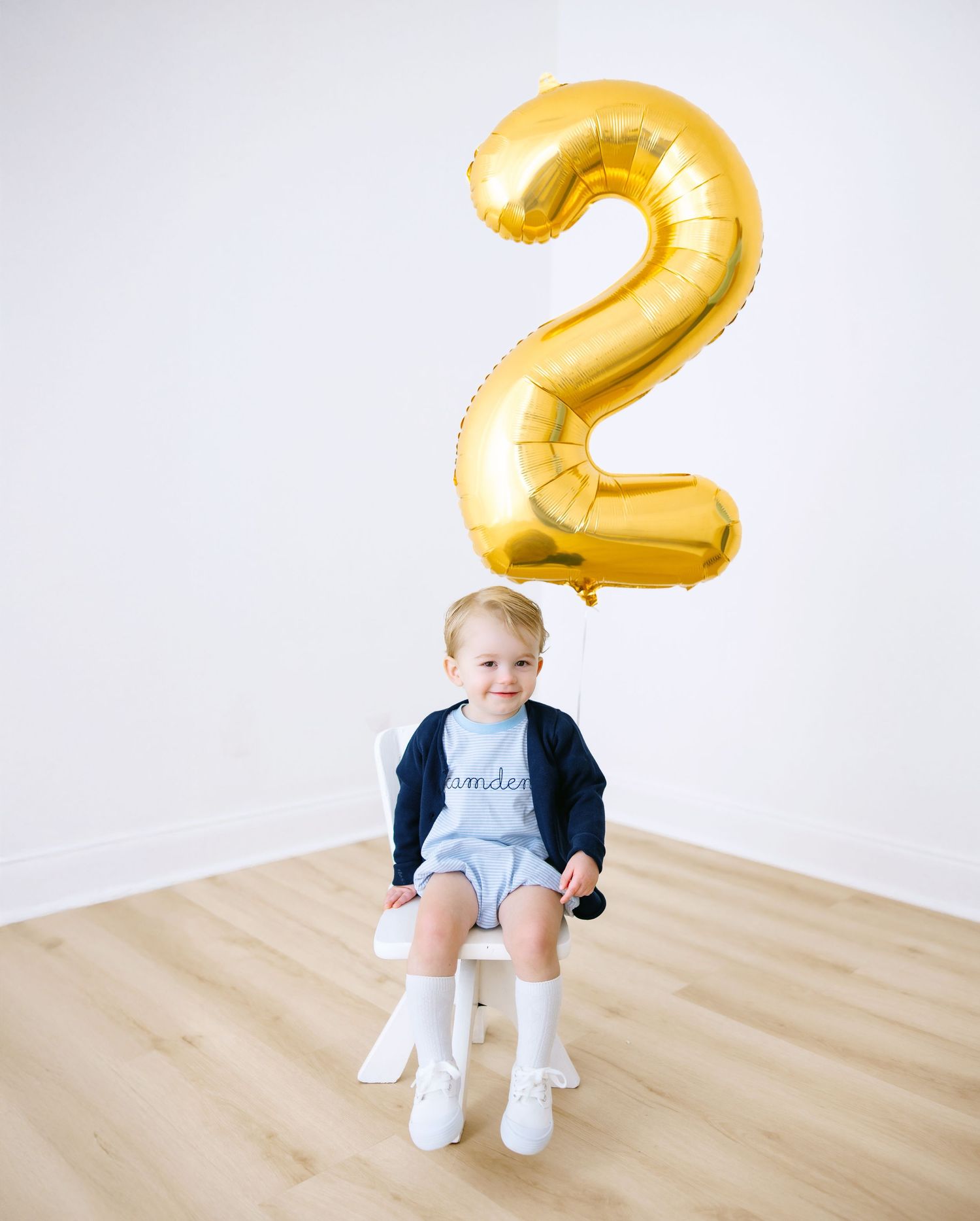 Small child sitting below a large gold number 2 balloon against a white backdrop for birthday photos.