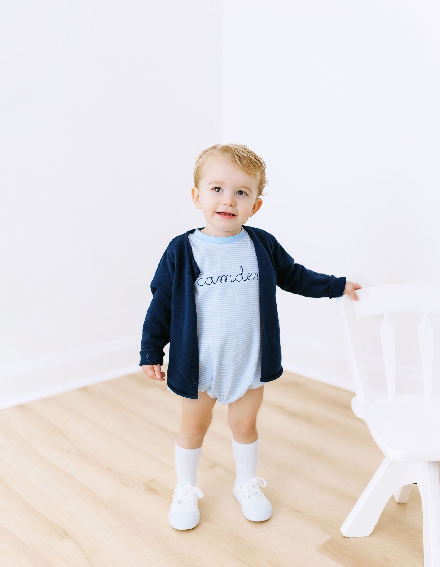 Small toddler wearing navy cardigan over light blue outfit with white socks and shoes on hardwood floor.