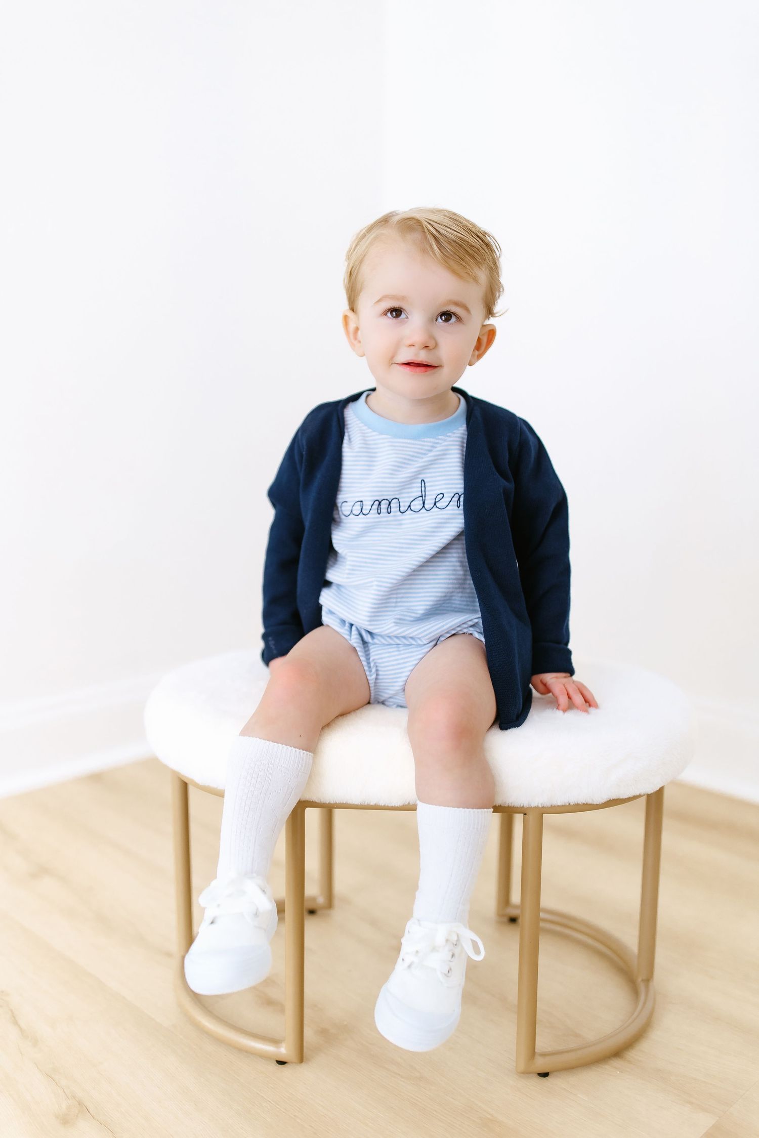 Small child sitting on white ottoman wearing light blue romper, navy cardigan, white socks and shoes against white background.