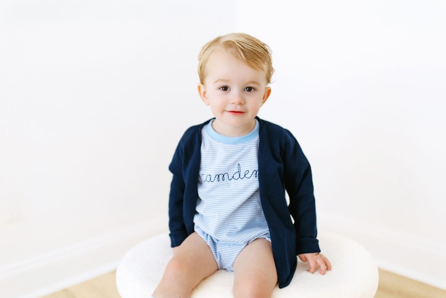 A small child wearing a light blue onesie and navy cardigan sits against a white backdrop.
