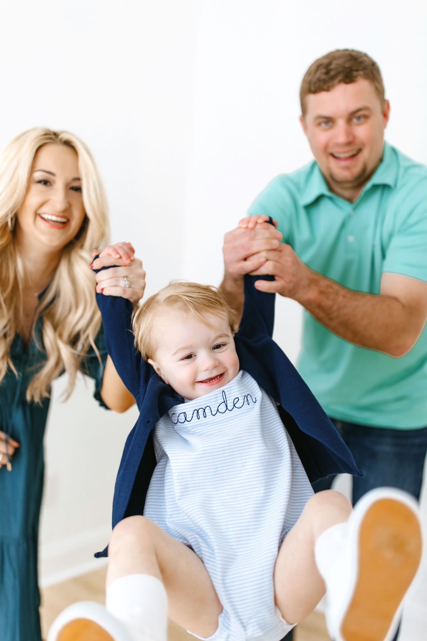 A happy family plays together as they lift a smiling toddler wearing a blue and white striped outfit up in the air.