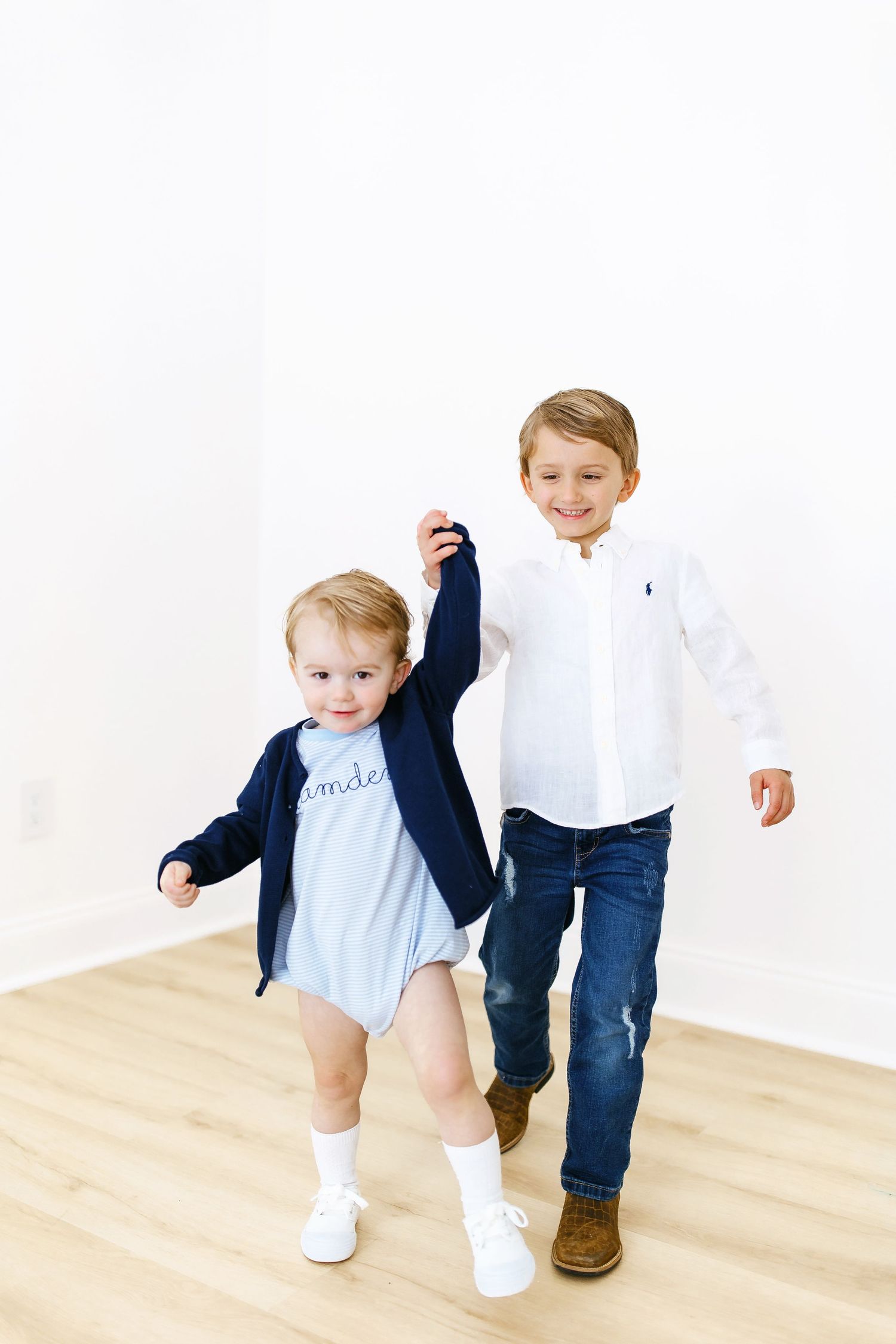 Two young children walking together on hardwood floors wearing navy and white clothing.
