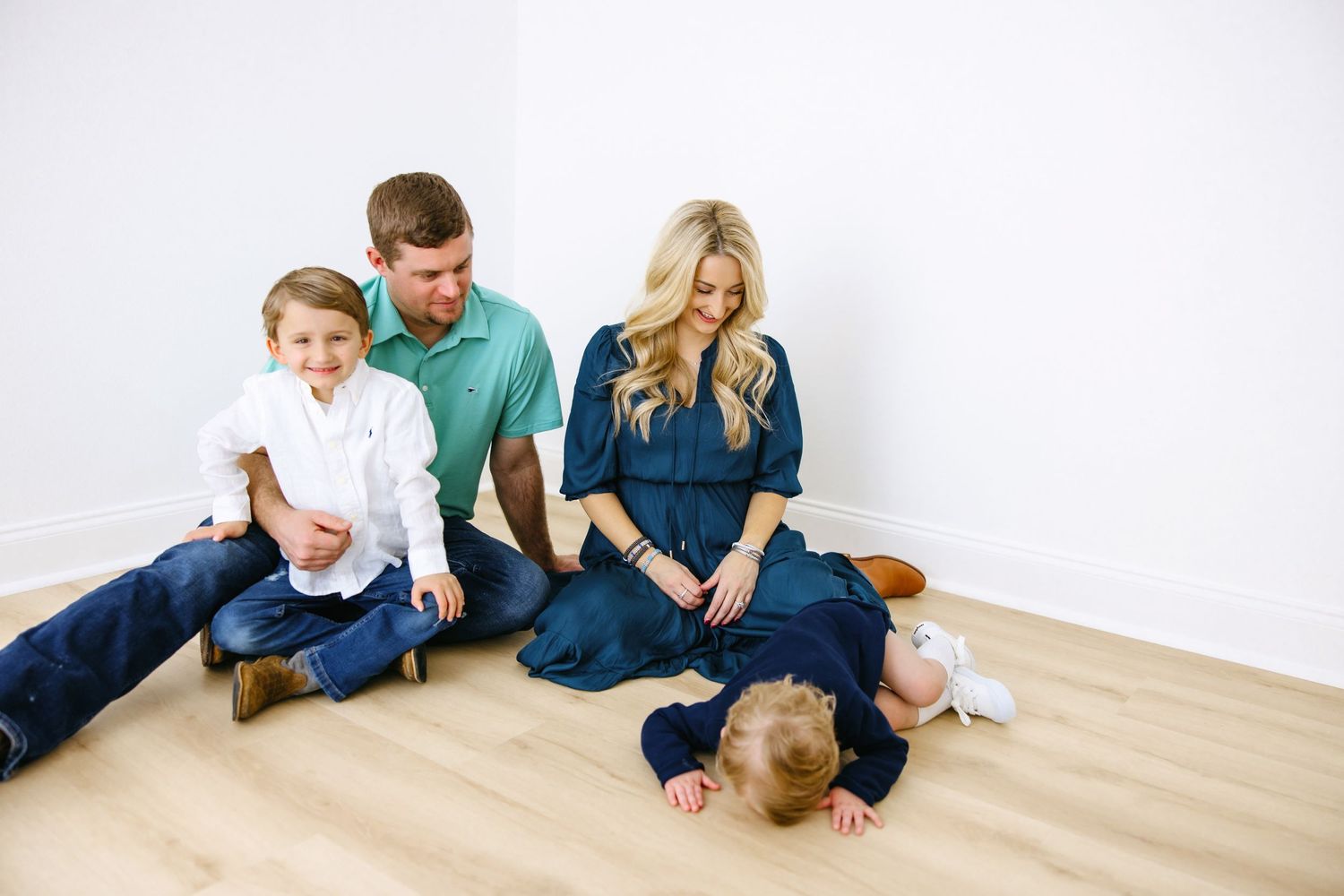 A family sits together on hardwood flooring against a white wall in casual clothing.