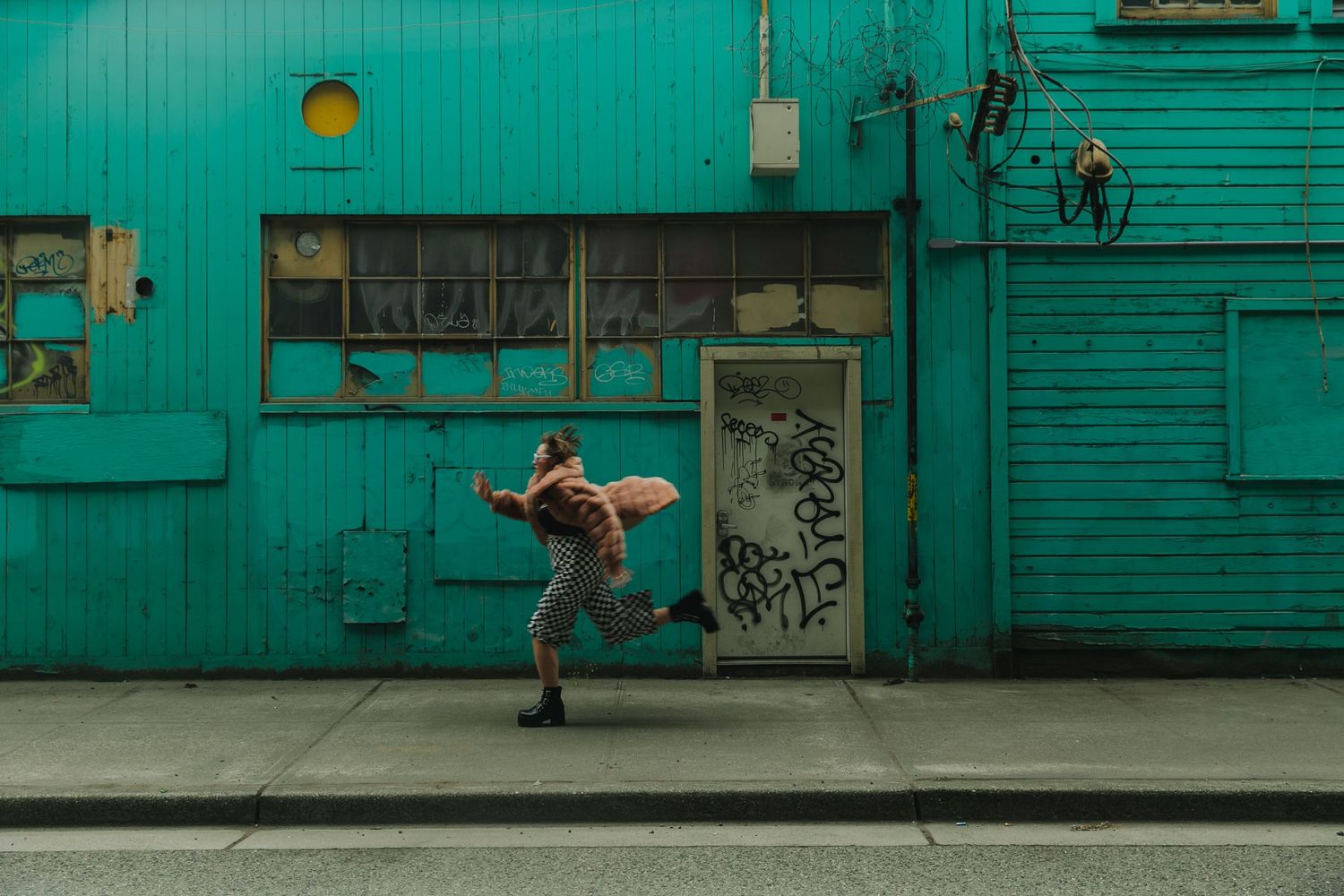 Person dancing energetically against vibrant turquoise wall with graffiti in an urban street setting.