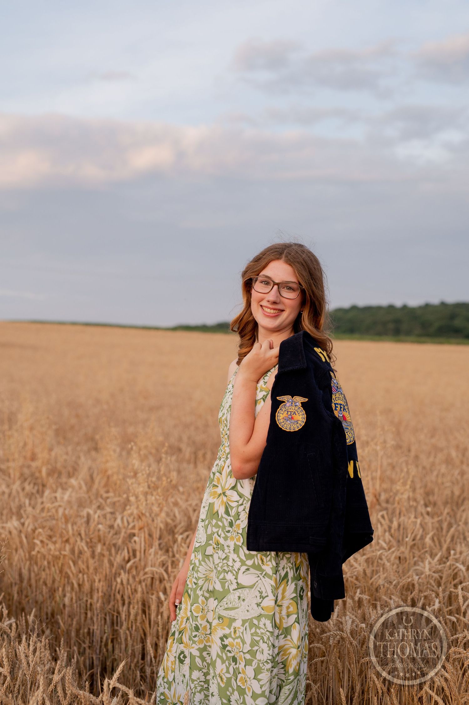 Someone wearing glasses and FFA jacket smiles while posing in a wheat field at sunset.