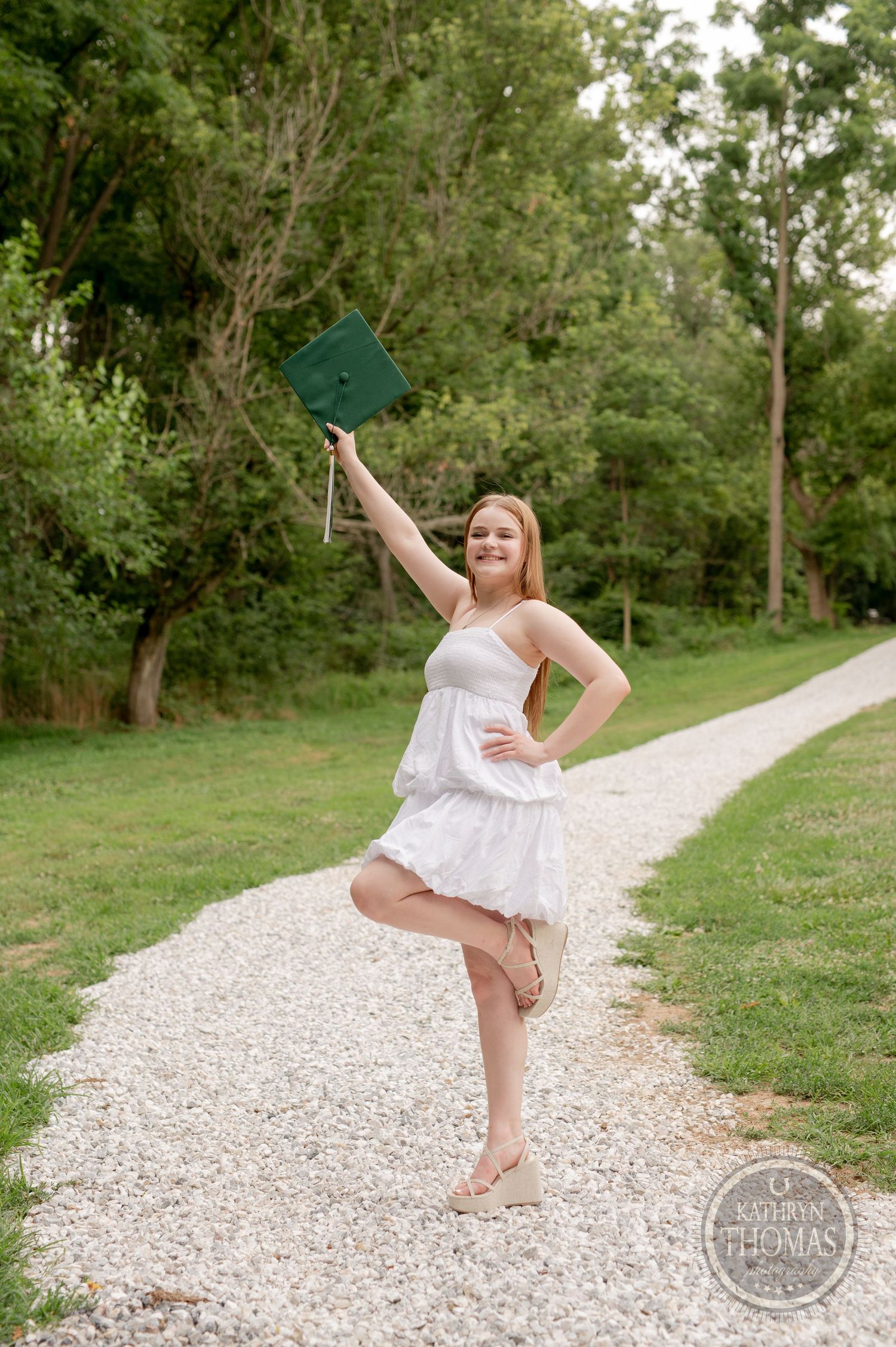 Graduate celebrating on gravel path holds diploma in the air while striking playful pose.