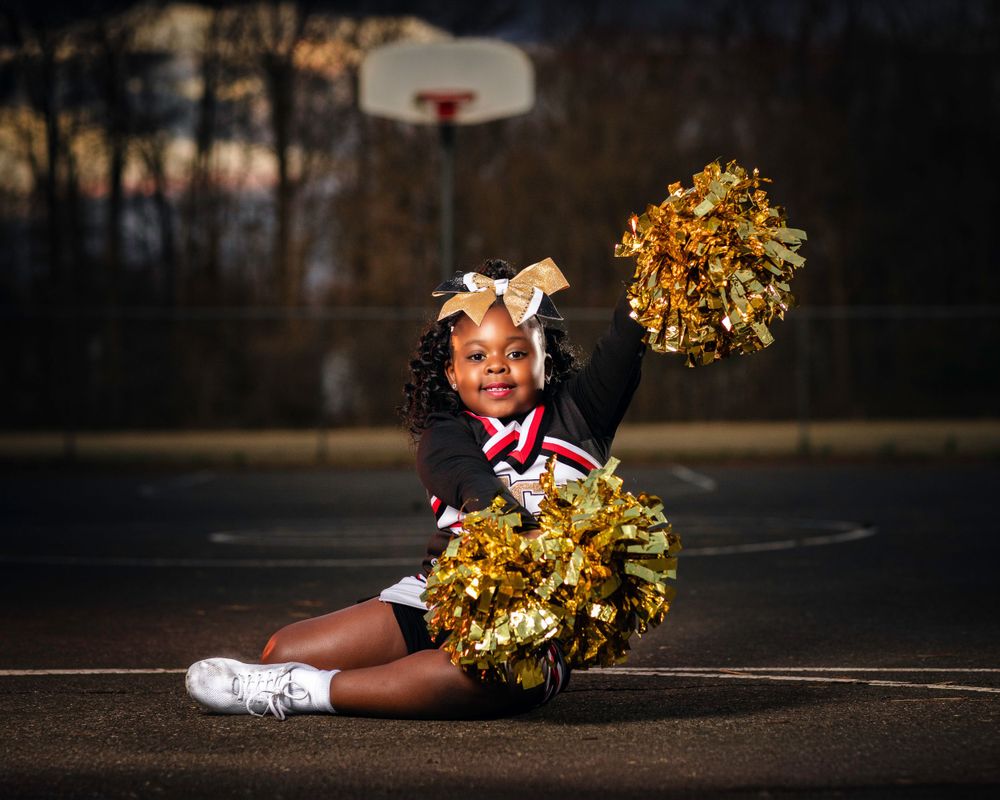Young cheerleader poses with gold pom poms on basketball court at dusk.