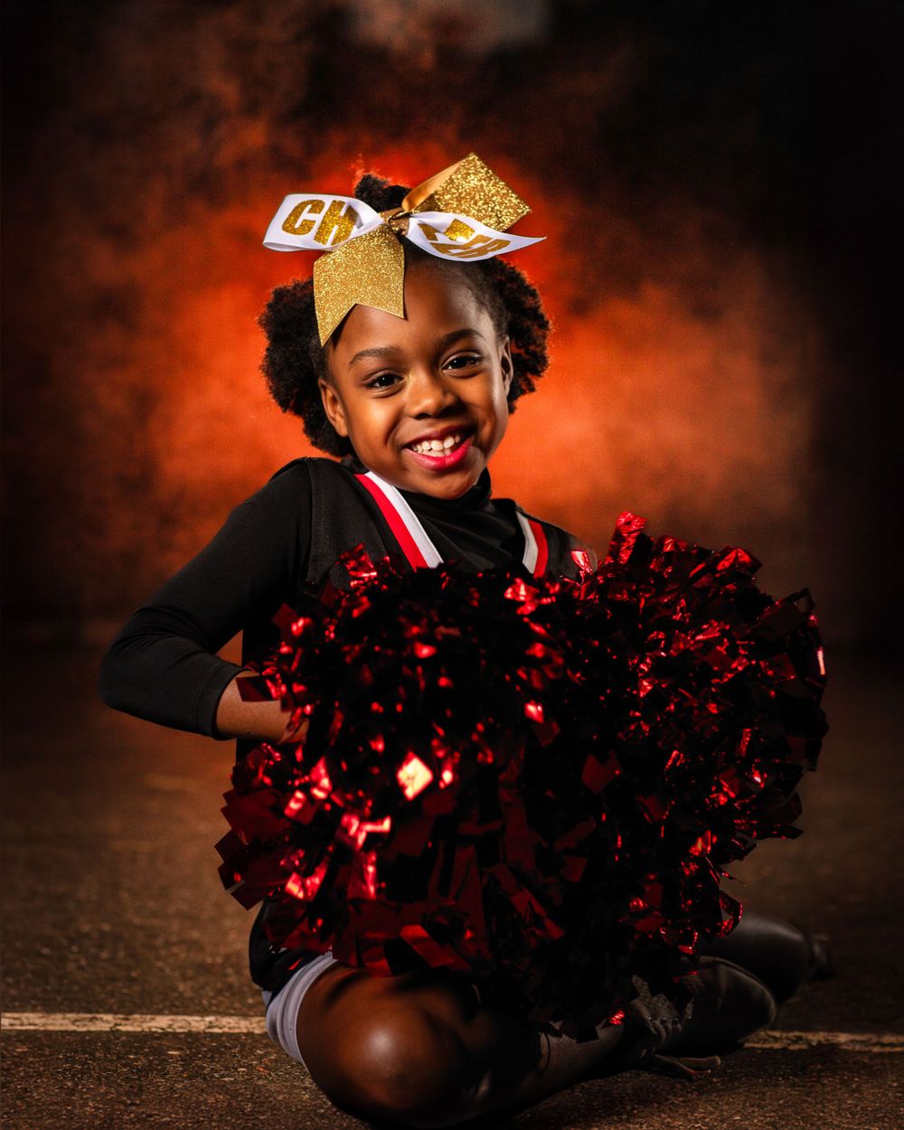 Cheerleader with red pom poms poses playfully on dark background with orange lighting.