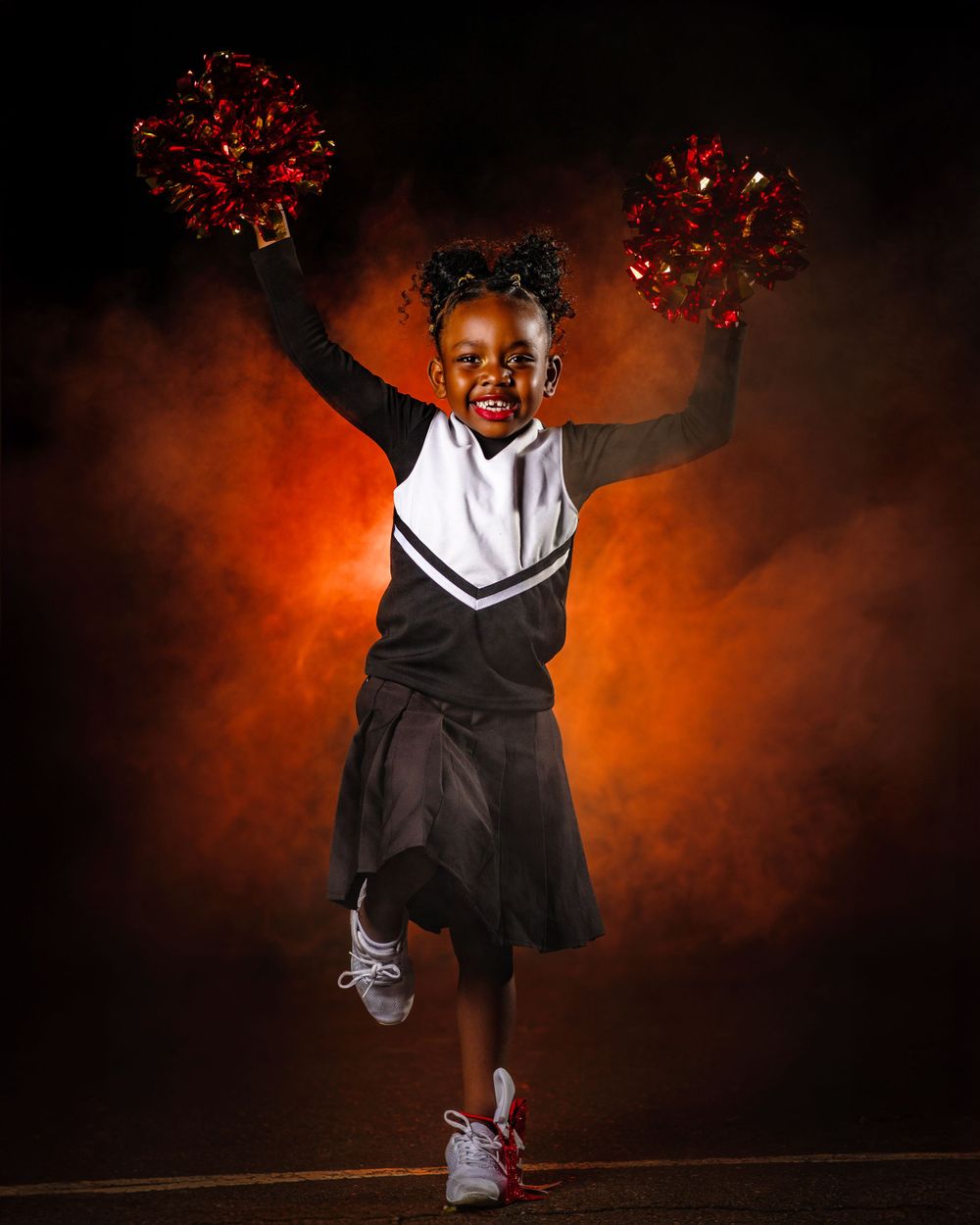 Young cheerleader in black uniform performs energetic dance moves against dramatic orange backdrop.