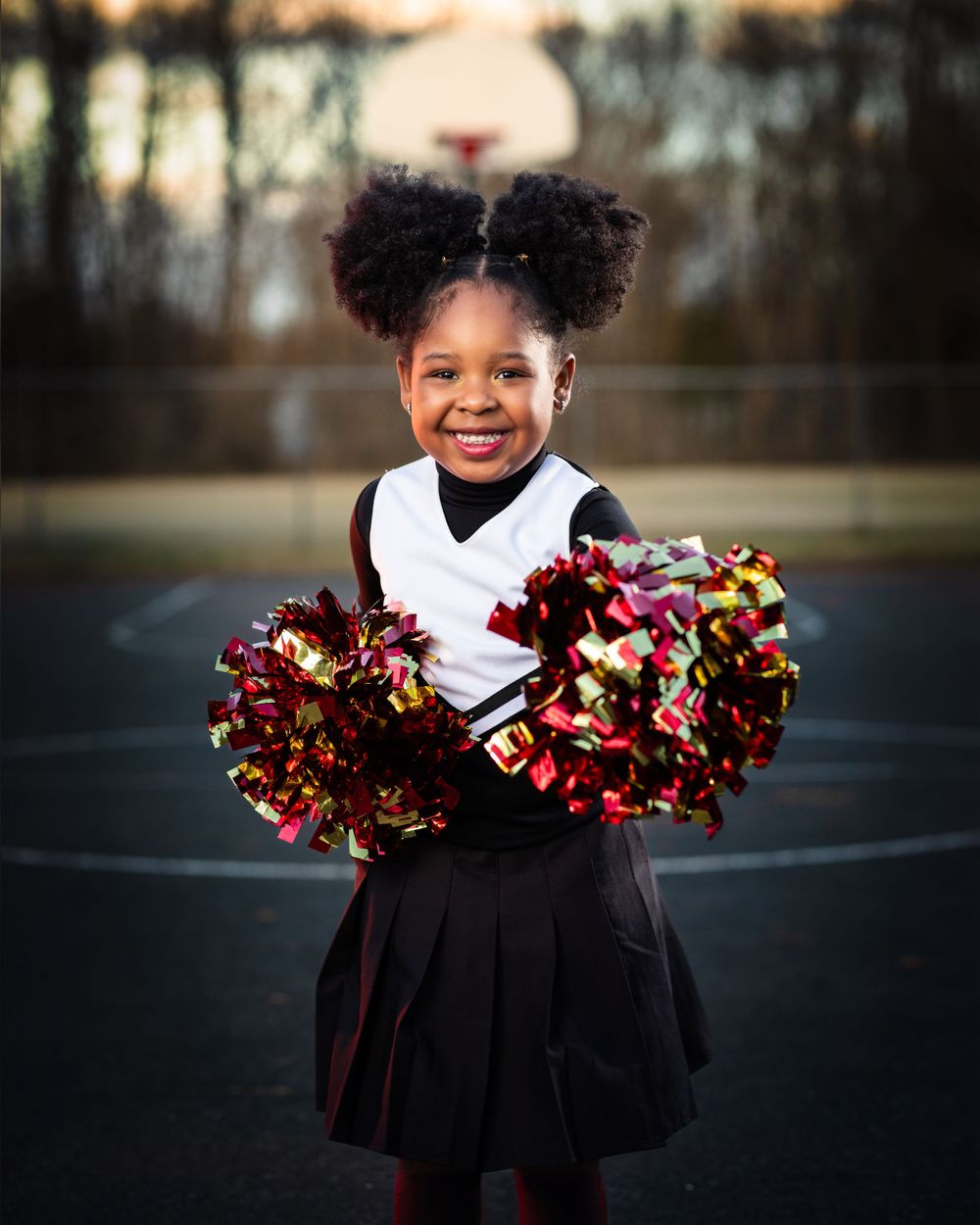 Cheerleader performs with red and gold pom poms on outdoor basketball court at sunset.
