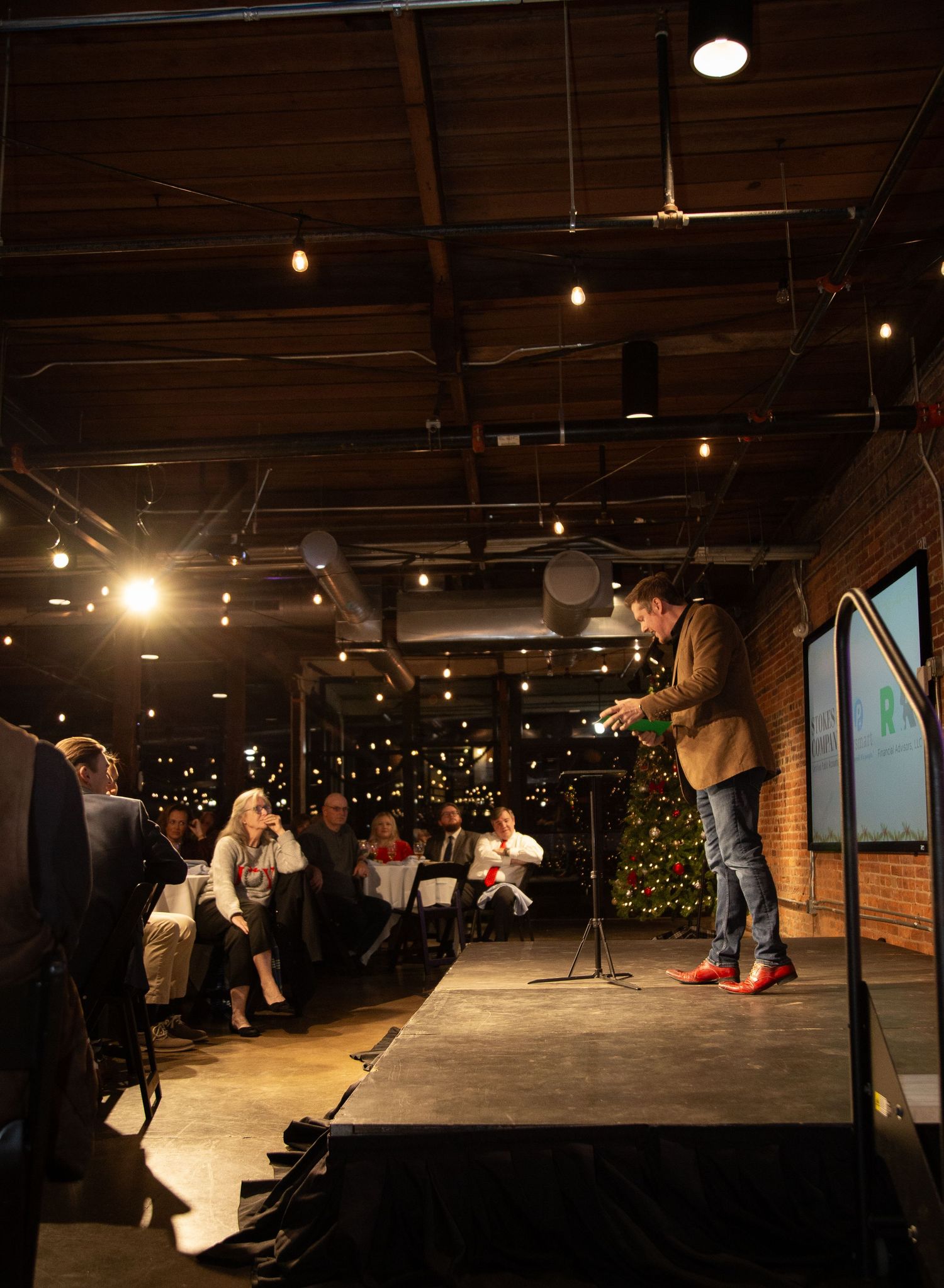 A performer, Bryan Saint, stands on a small stage in a dimly lit venue with exposed brick walls and hanging lights above the audience.
