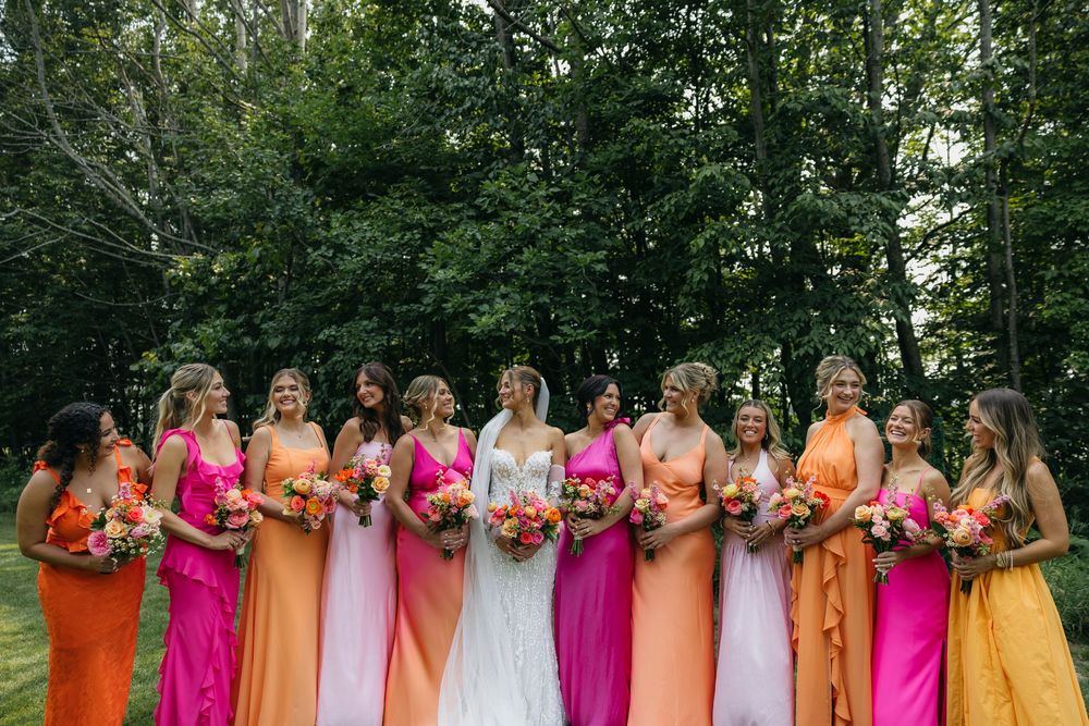 Bridesmaids in vibrant rainbow colored dresses stand together holding bright floral bouquets outdoors.