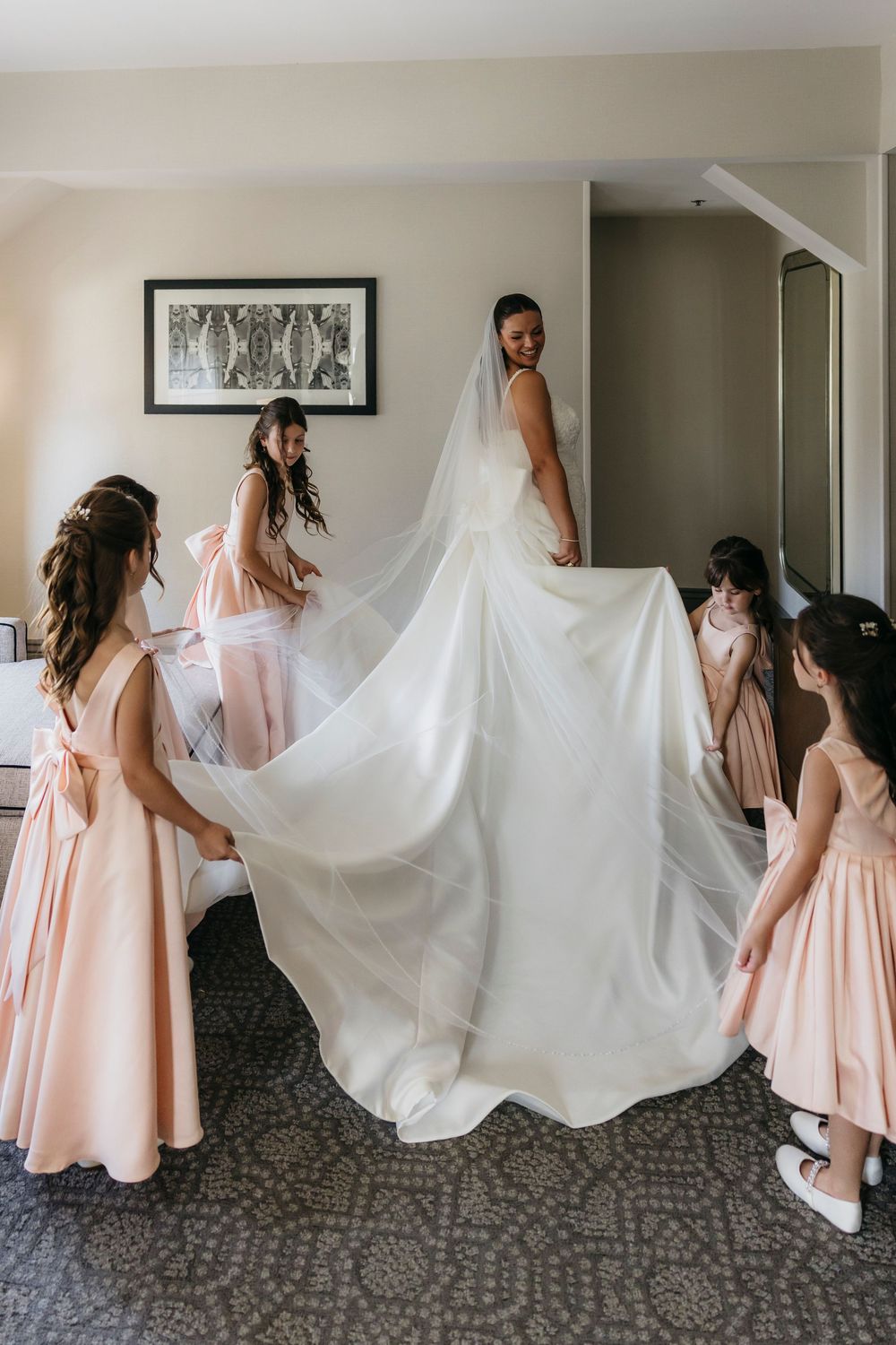 Bridesmaids in blush pink dresses help with a flowing white wedding dress train in a hotel room.