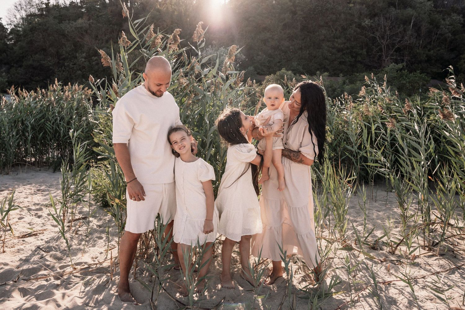 Happy family in white outfits posing together among tall grass at sunset.