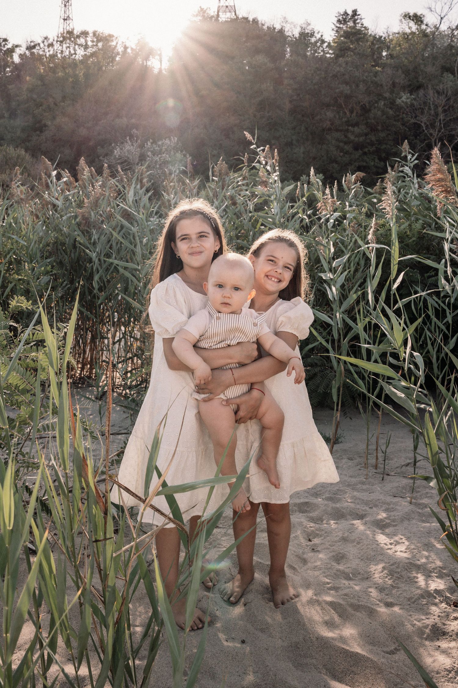 Three people in white dresses pose among tall reeds at sunset.