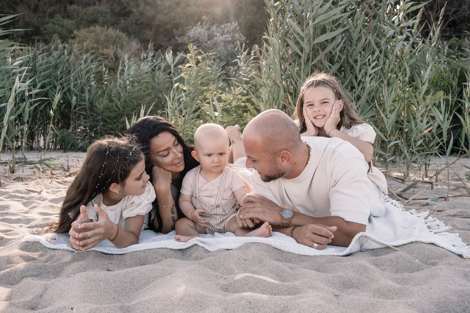 A family lounges together on a sandy beach during sunset wearing coordinated white outfits.
