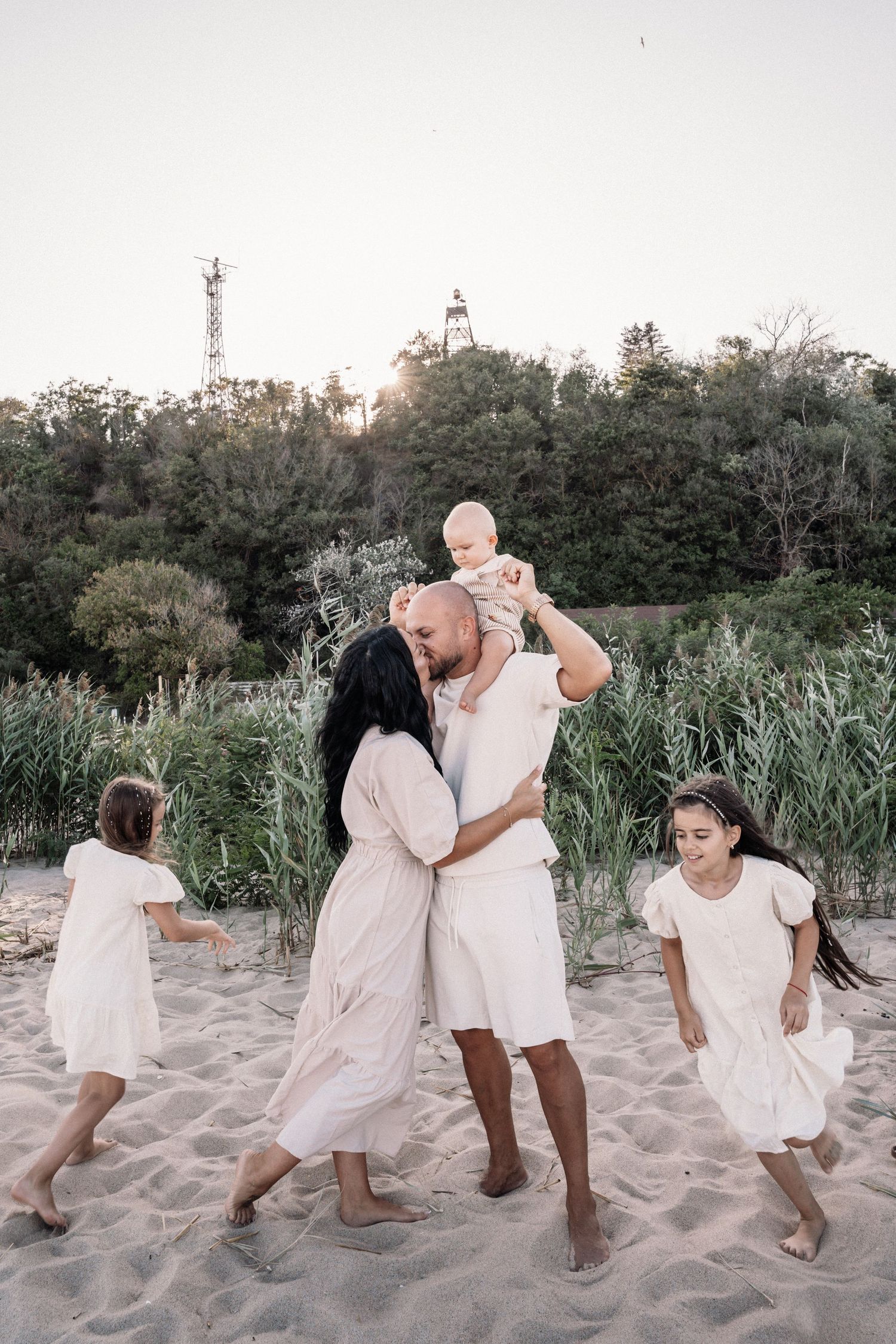 Playful family dances together on beach sand wearing matching white clothing at sunset.