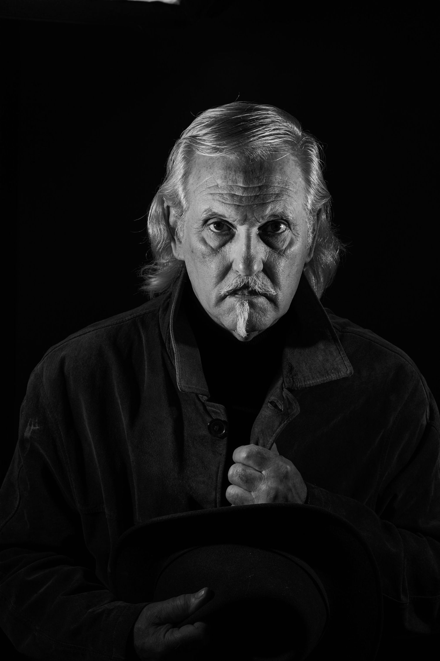 Dramatic black and white portrait of a person in dark clothing against a black background with an intense expression.