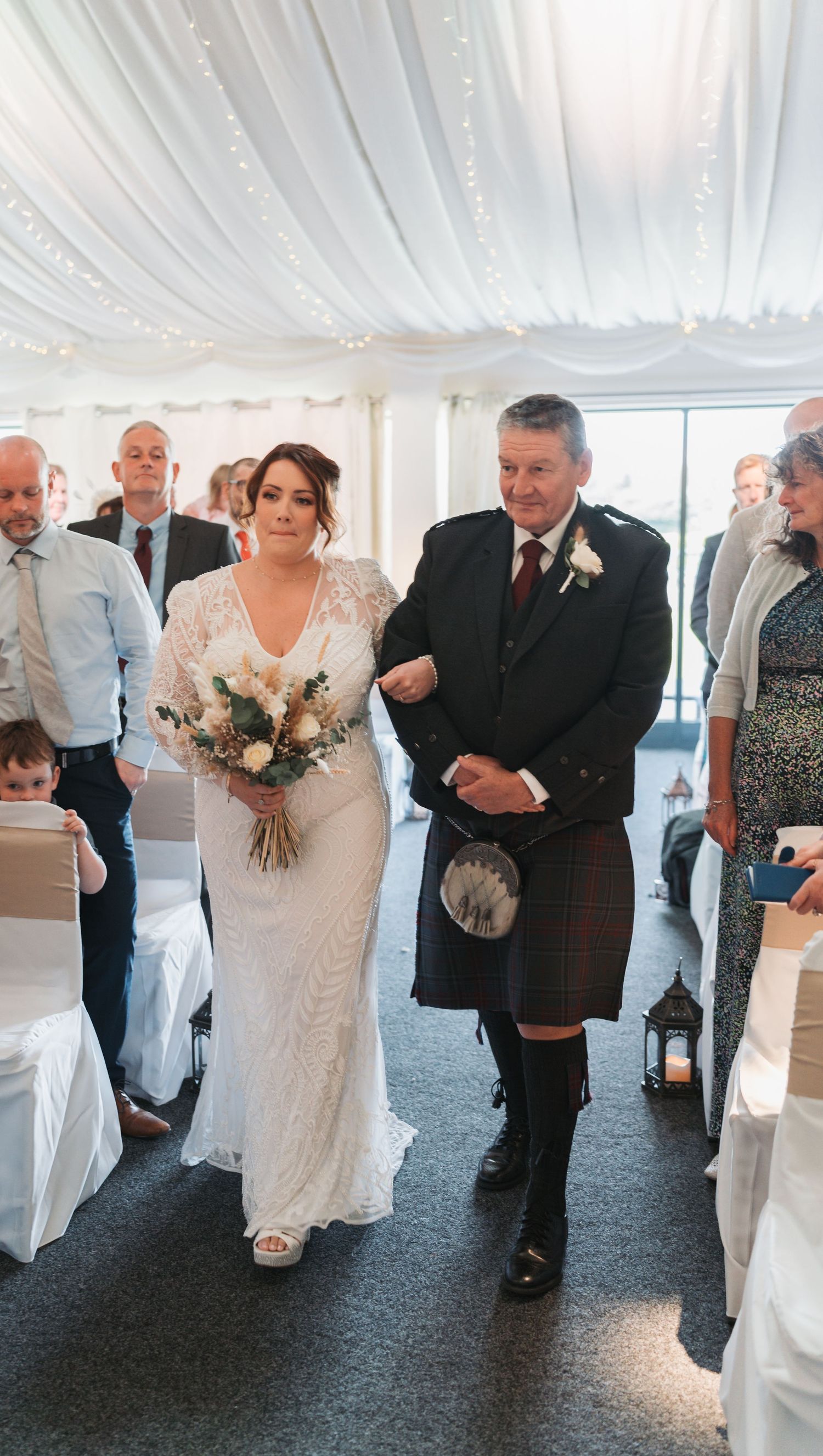 A wedding ceremony in a white draped tent with people in formal attire including Scottish kilts and elegant dresses.