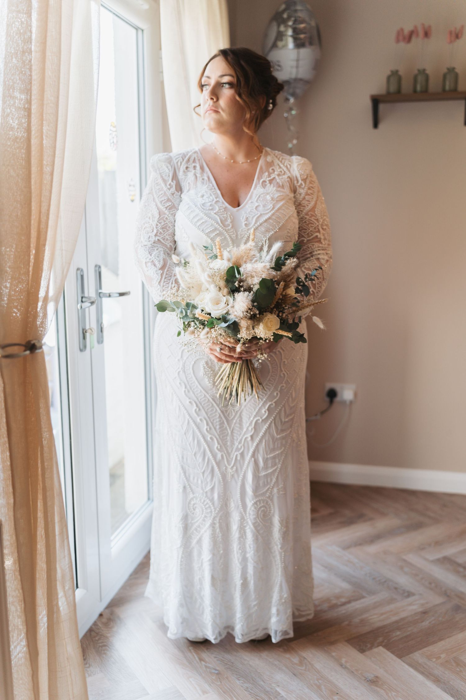 Elegant lace wedding dress with long sleeves and detailed embroidery stands near window holding white floral bouquet.