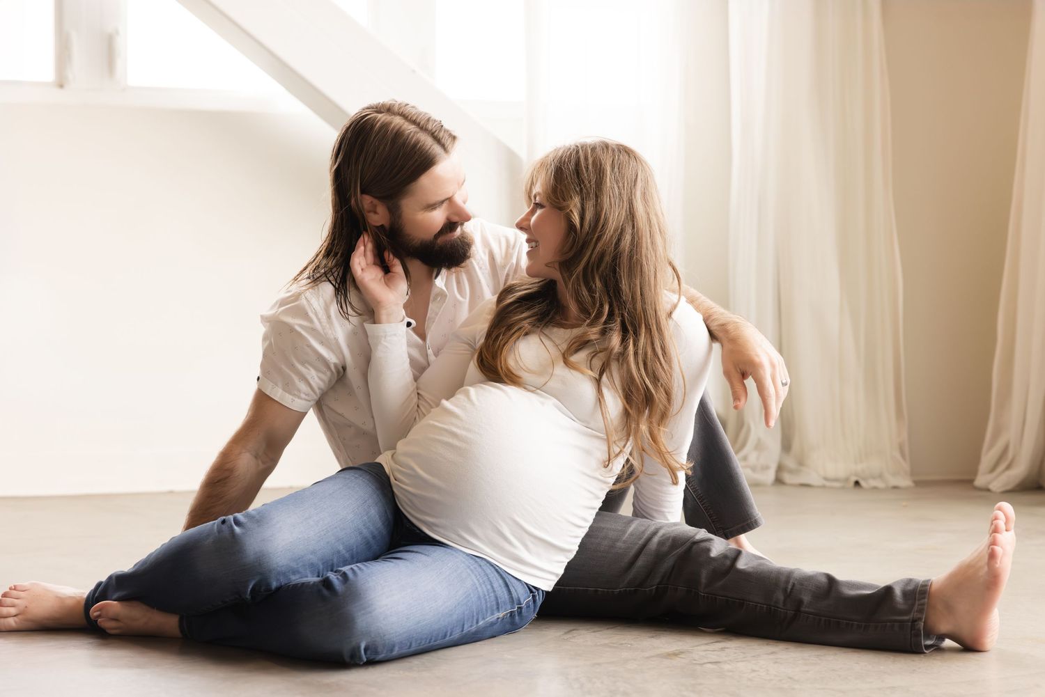 Happy couple in casual clothing sitting on floor by window in bright room sharing intimate moment together.
