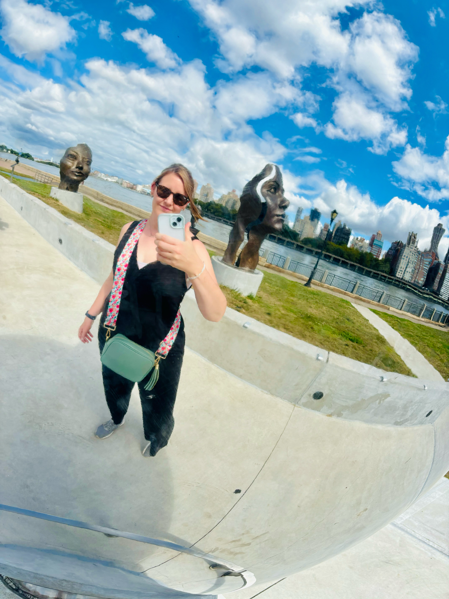 A woman takes a mirror selfie at an outdoor sculpture park with large face sculptures and blue sky.