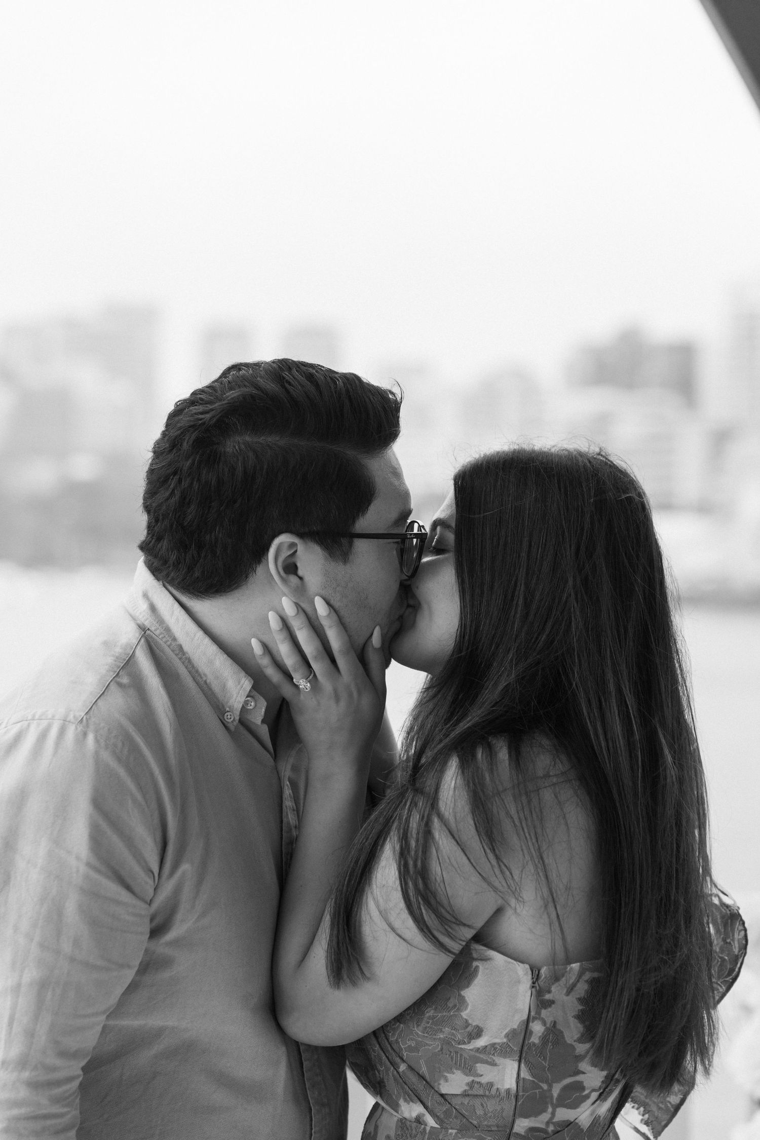Intimate black and white portrait of a couple sharing a romantic kiss with city skyline backdrop.