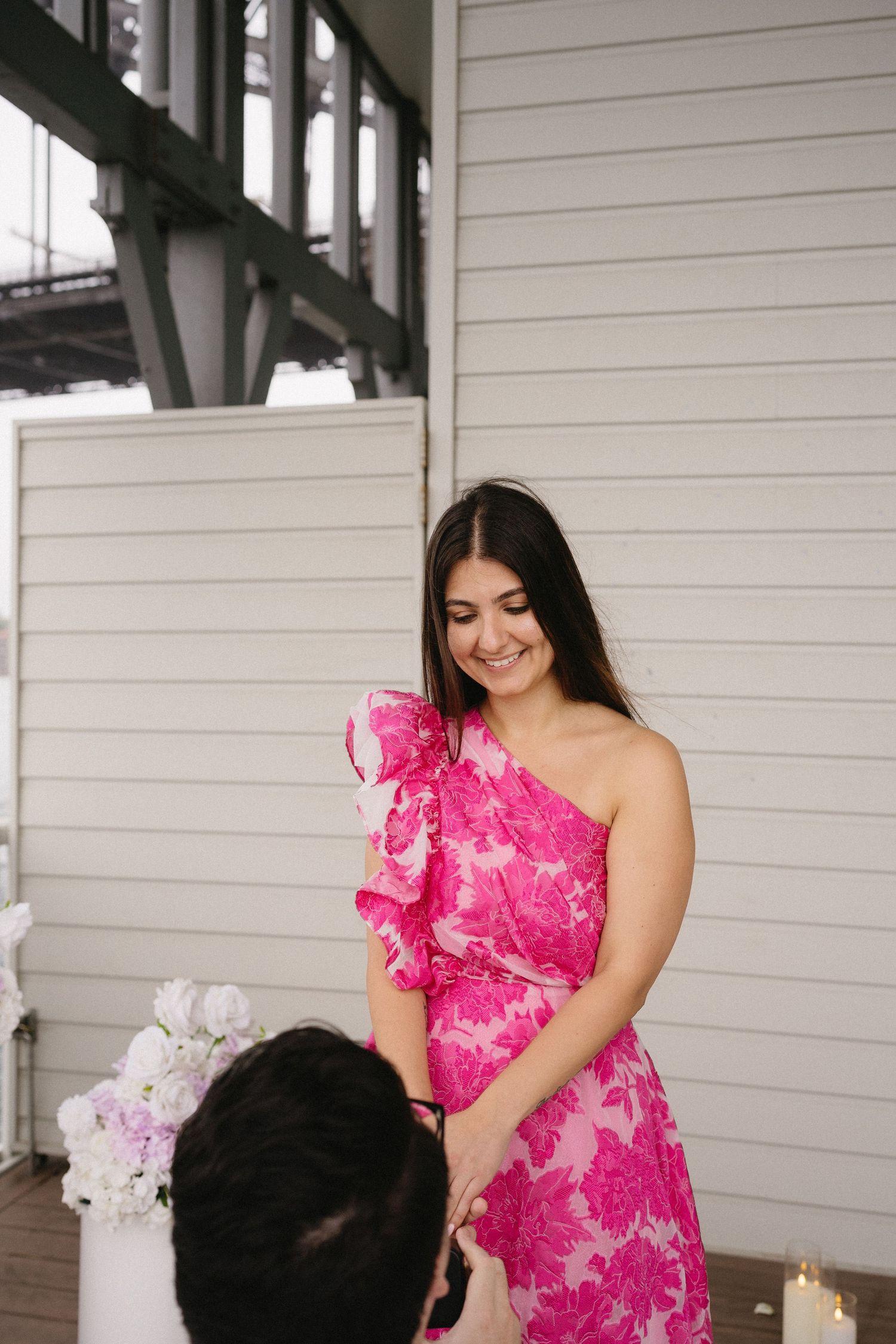 Woman in stunning pink one-shoulder dress smiling during romantic moment on porch.