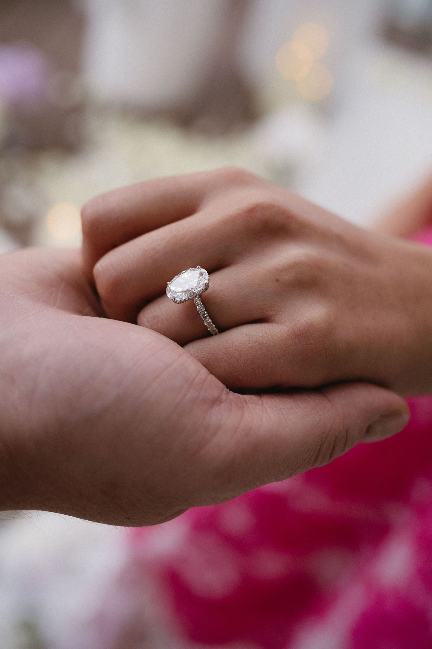 Close-up of elegant oval engagement ring being placed on finger with pink dress visible.