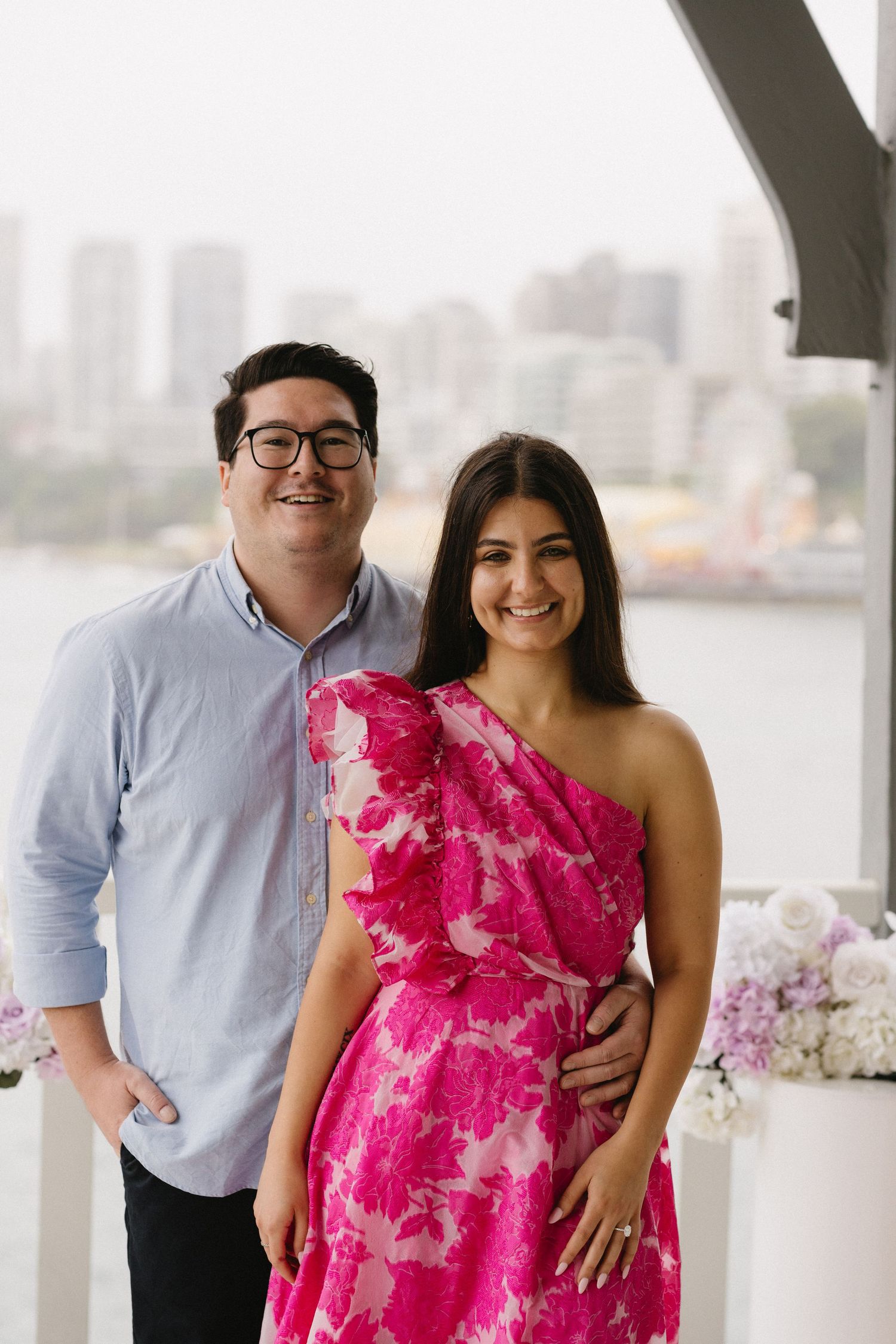 Couple in coordinated outfits posing on balcony with city views, woman wearing vibrant pink floral dress.