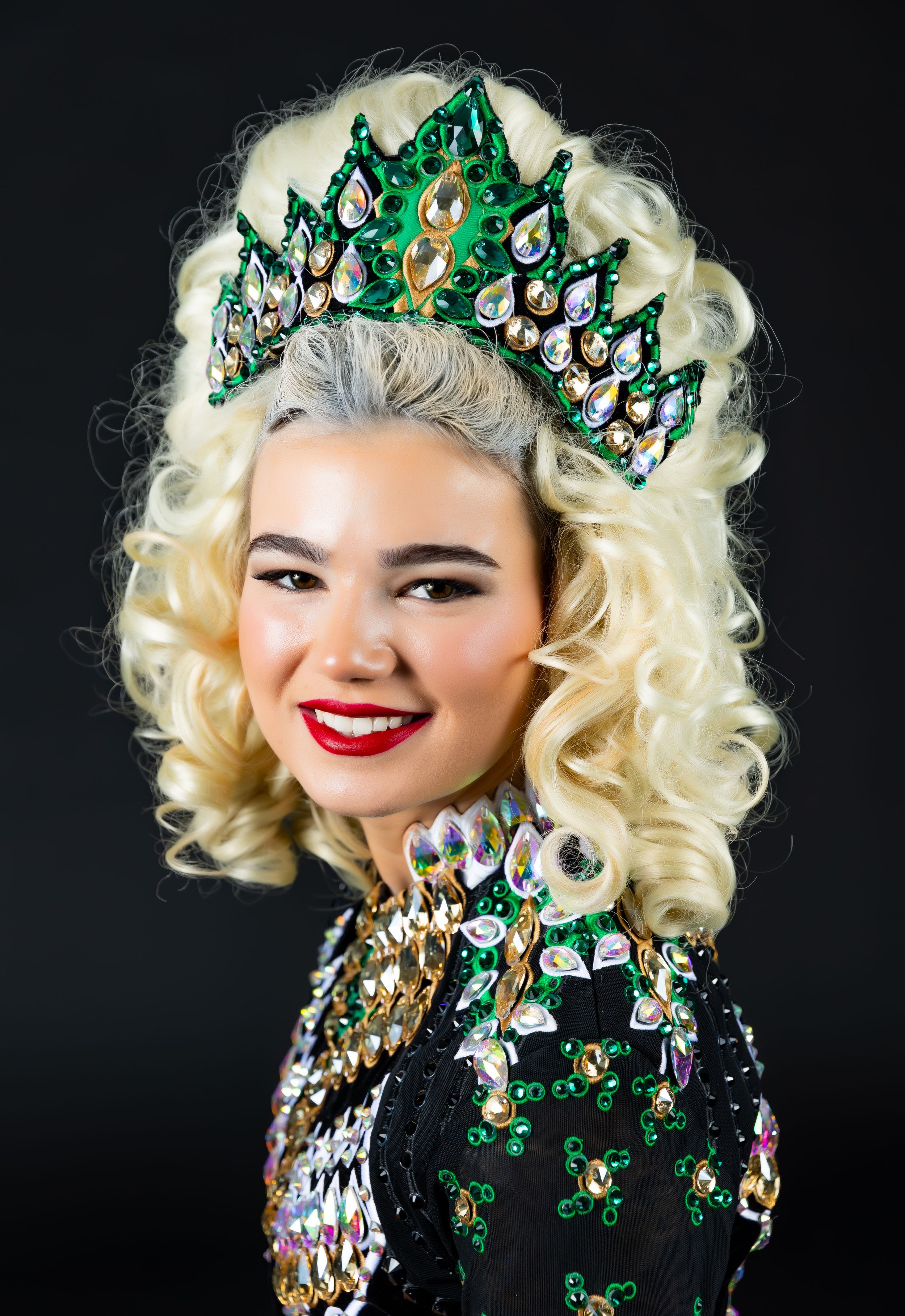 A performer wears a stunning emerald green and crystal crown with blonde curls and a sequined costume against a dark background.