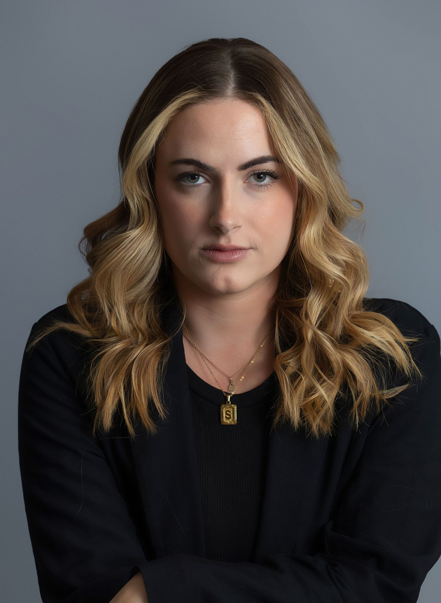 Professional headshot against grey background featuring wavy blonde hair and black outfit.