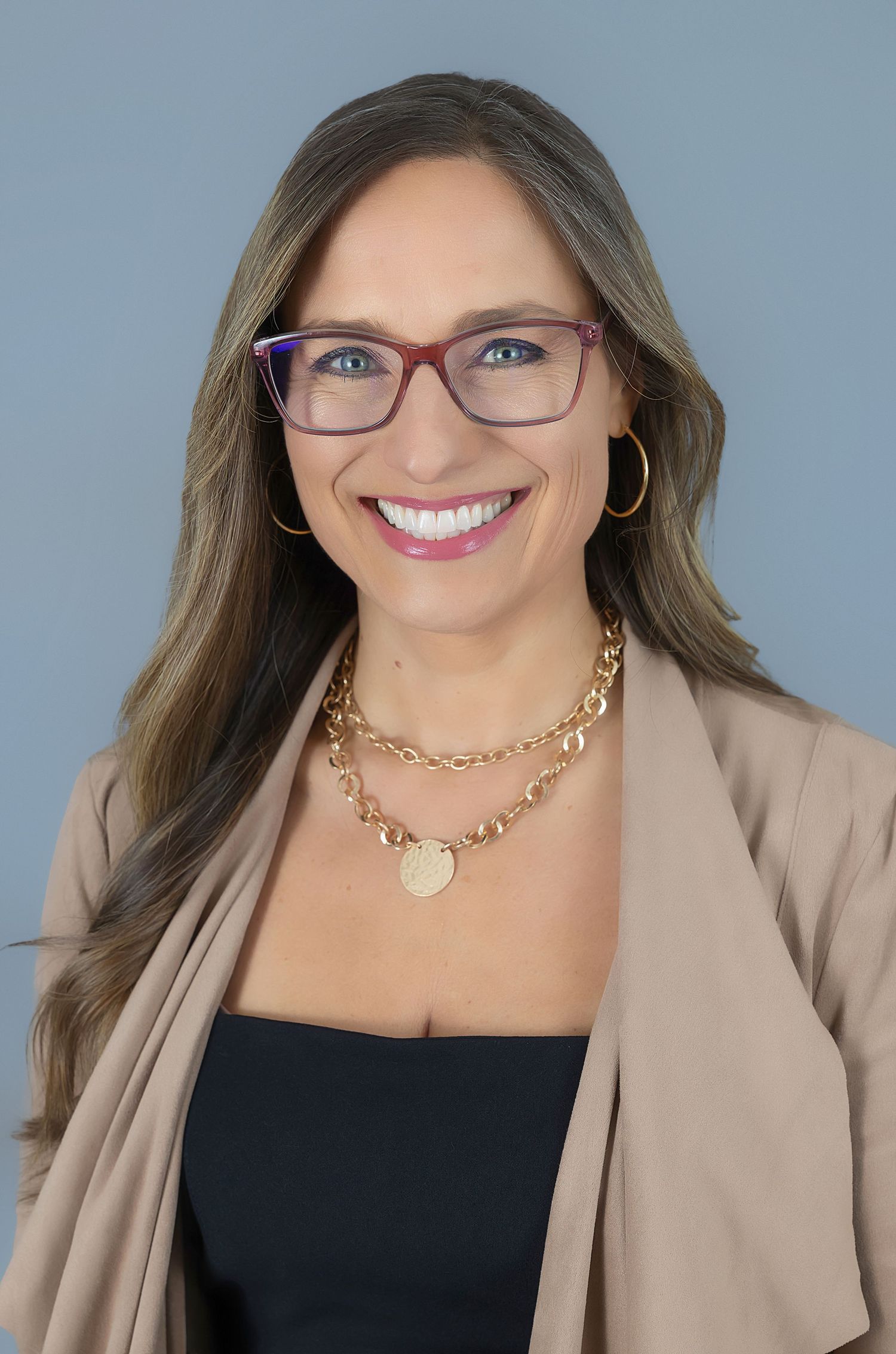 Professional headshot of person in business attire wearing red glasses and layered gold necklace.