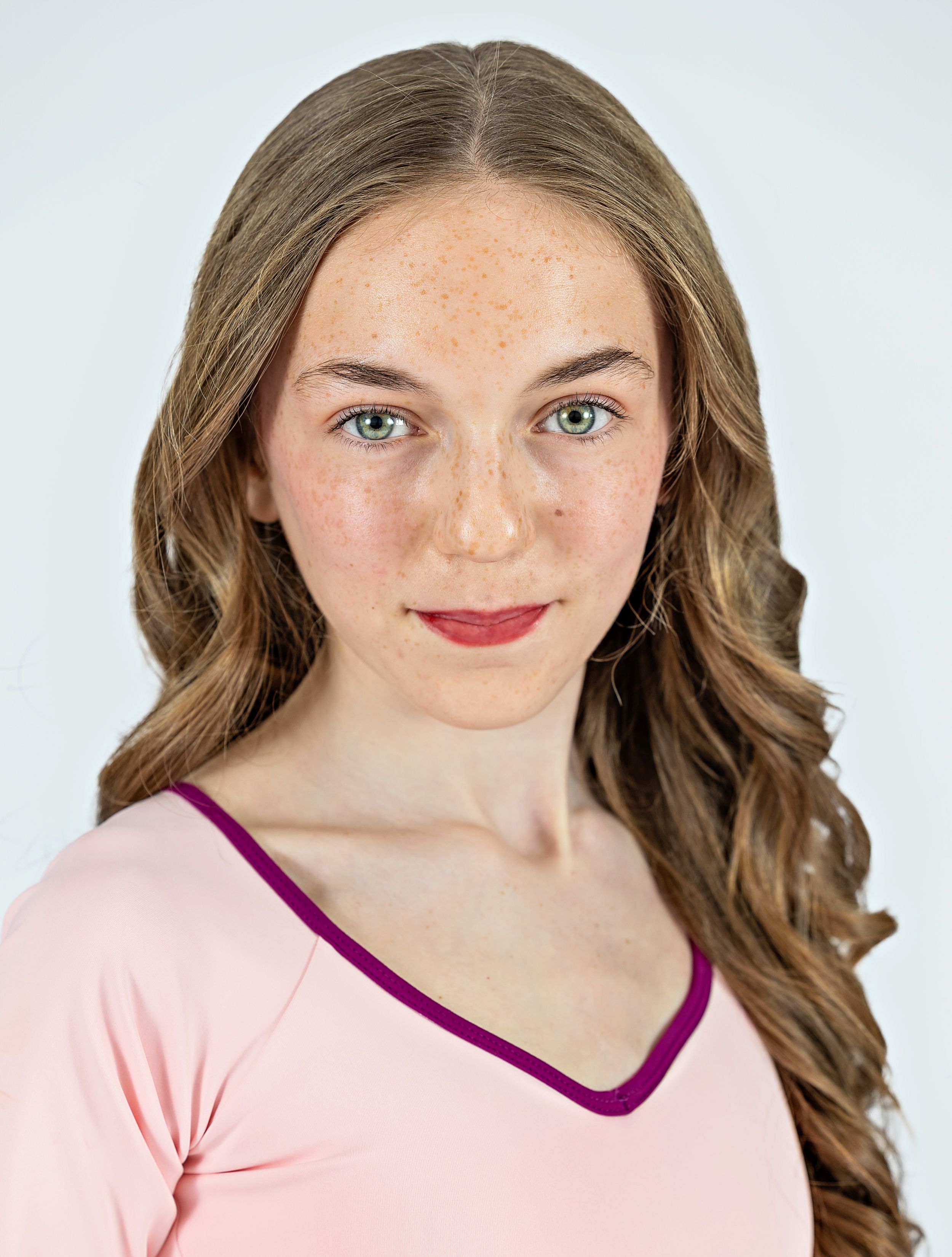 Portrait against white background wearing pink v-neck top with burgundy trim and long wavy brown hair.