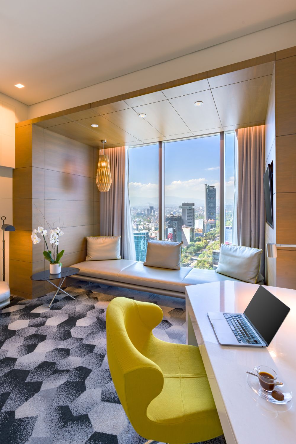 Modern office space with window seat, yellow accent chair, and laptop desk overlooking city skyline through large windows.
