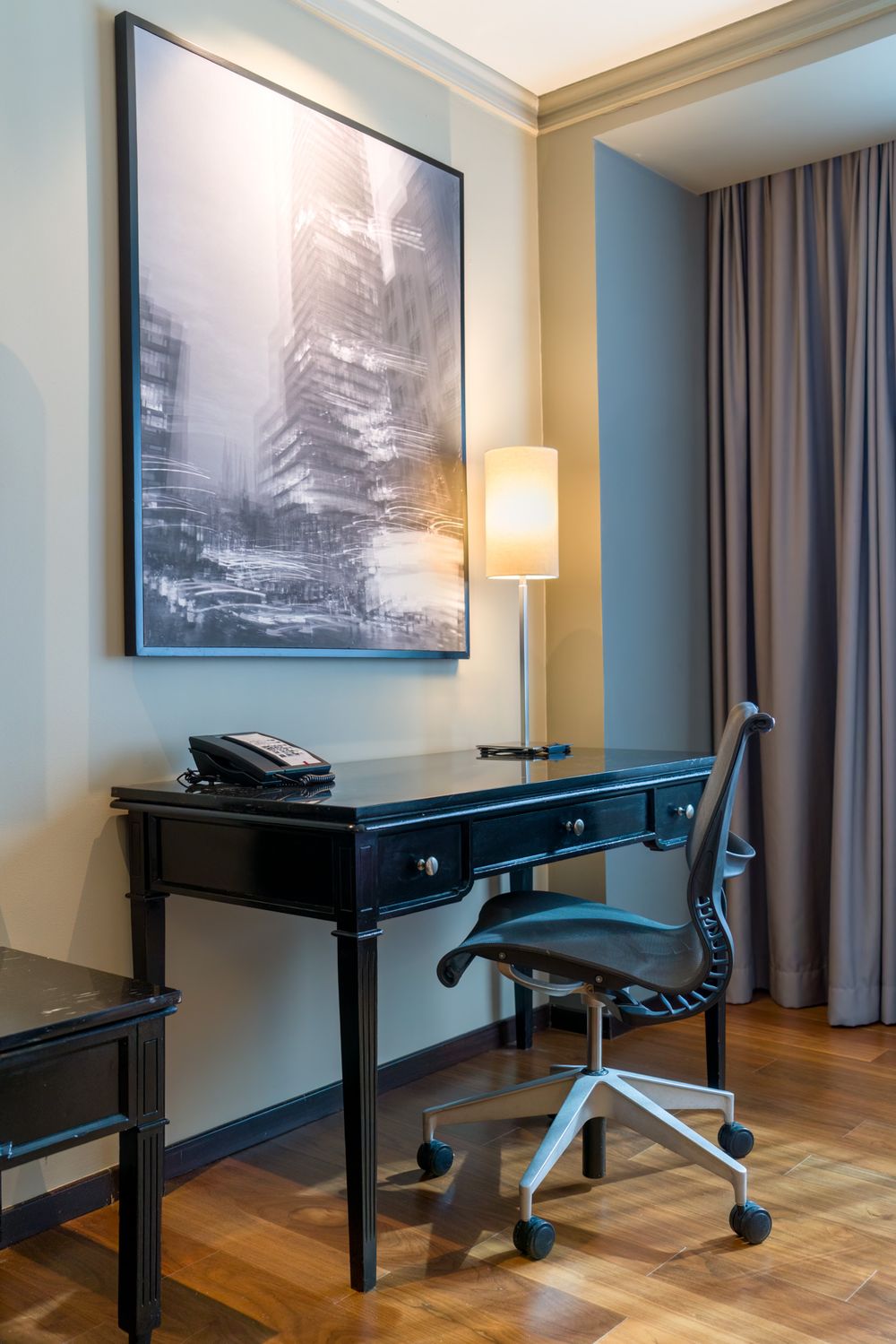 Modern hotel room workspace with black desk, ergonomic chair, and large monochromatic cityscape artwork on blue walls.