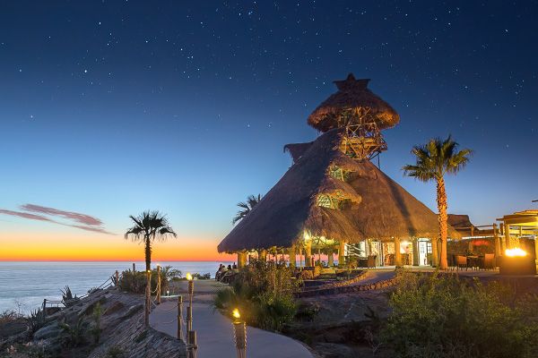 Thatched-roof resort restaurant overlooking ocean at sunset with palm trees and glowing lights. El Mirador by Guaycura.
