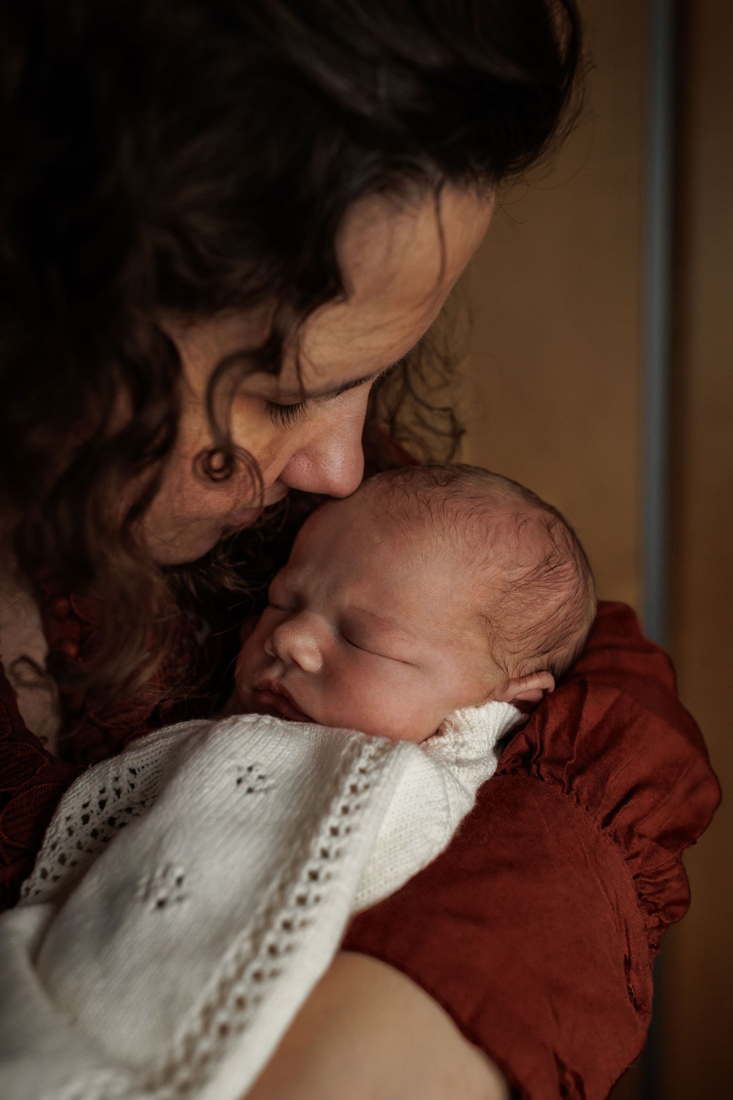 A tender moment between parent and newborn baby snuggling together in warm lighting, wrapped in white and red fabric.