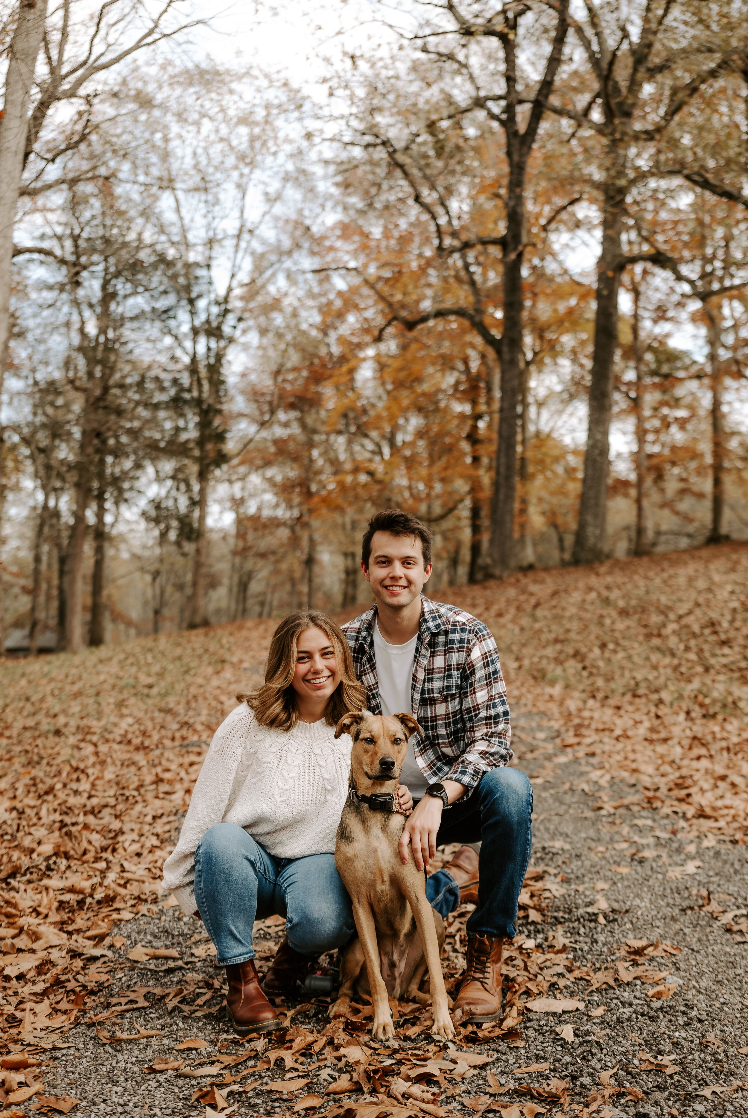 Couple and their brown dog sitting together on fallen autumn leaves in wooded area.