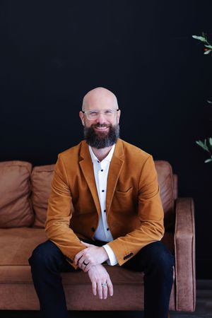 Person in tan blazer and white dress shirt sits on brown leather couch in studio setting with dark backdrop.