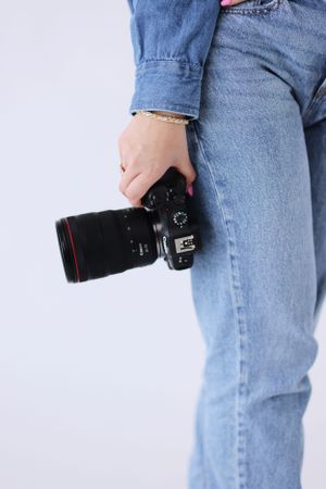 Person wearing blue jeans holds a black Canon DSLR camera with telephoto lens against white background.