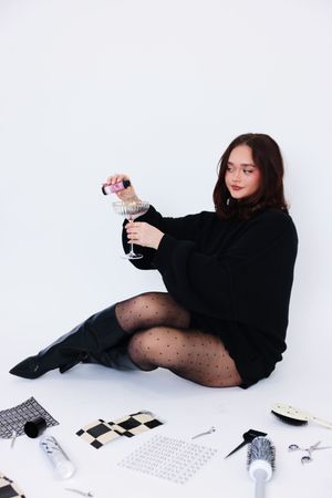 A person in black attire and polka dot tights lies on a white surface with scattered photos and a wine glass.