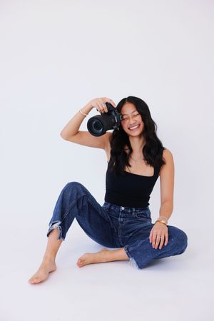 A photographer in a black top and denim jeans poses playfully on a white background while holding a camera.