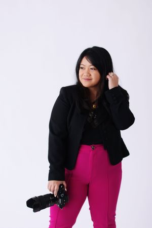 Professional photographer wearing black blazer and bright pink pants holds camera against white studio background.