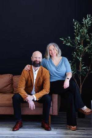 Professional studio portrait of two adults sitting on a brown leather couch with a tall plant against a dark background.