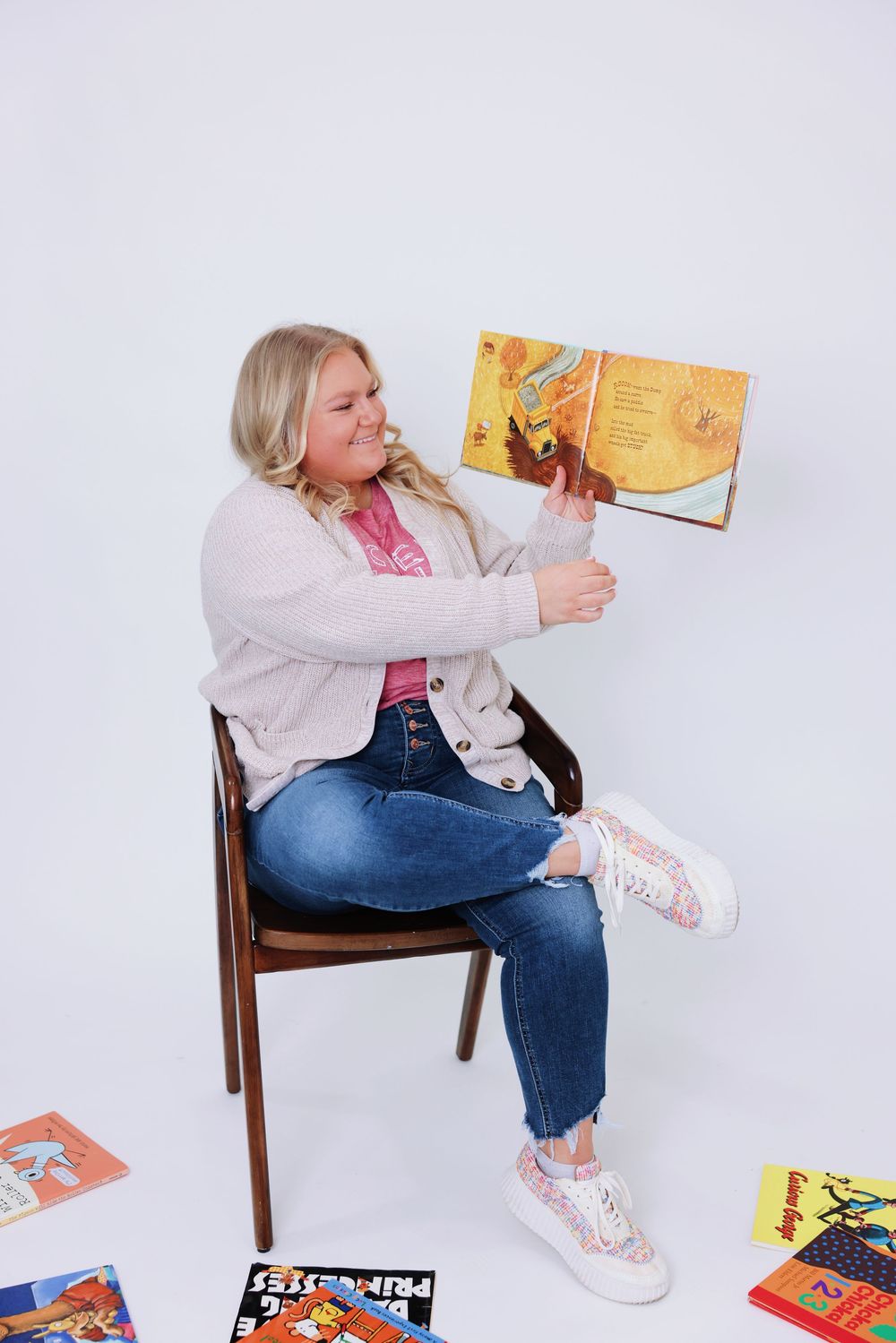 A person in jeans and white sneakers sits on a wooden chair reading colorful children's books scattered on the floor.